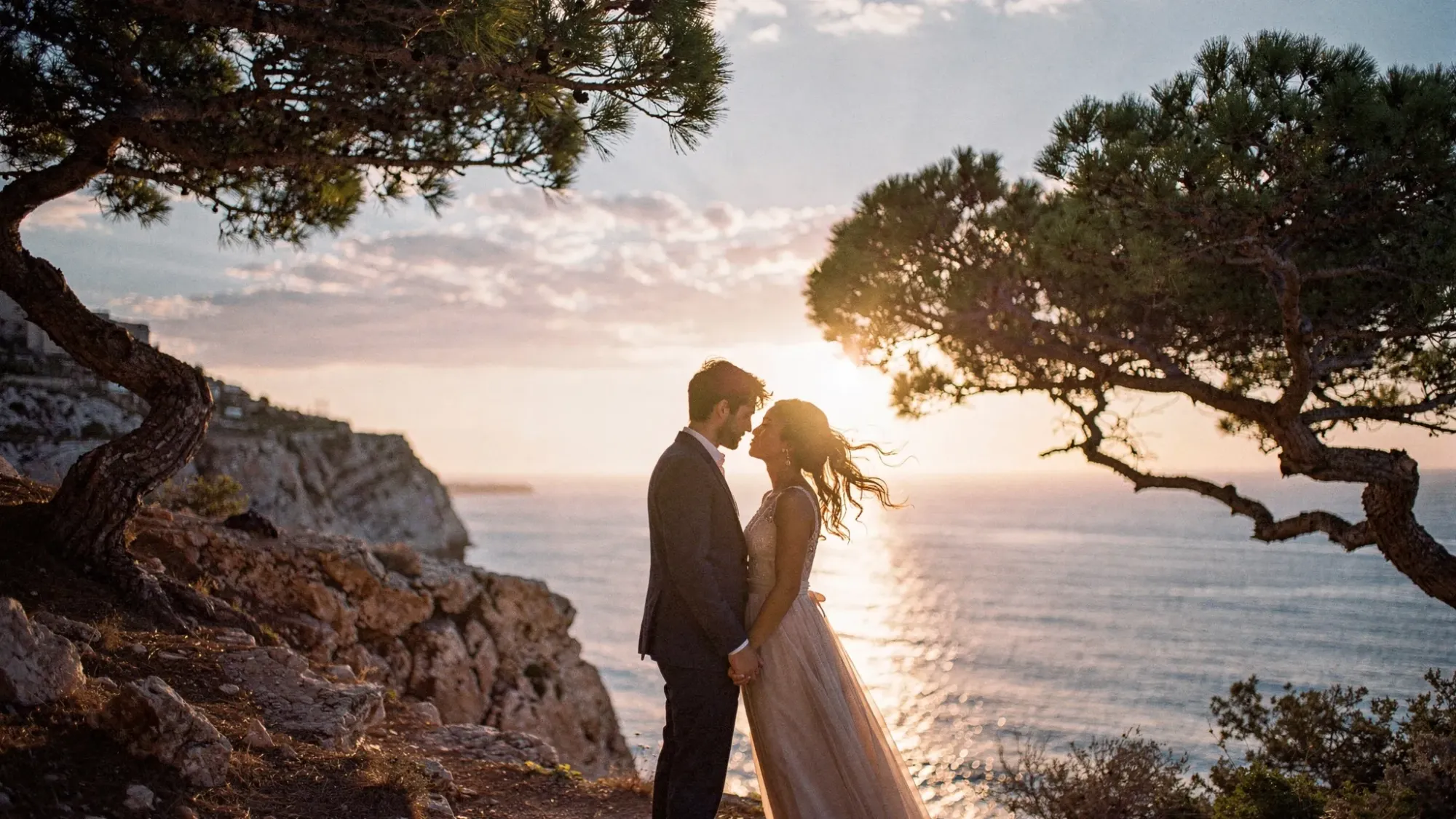 An intimate elopement moment on a Mediterranean sea cliff at sunrise, with two people standing close in the wind, pine trees nearby, and golden light spilling across the water below.