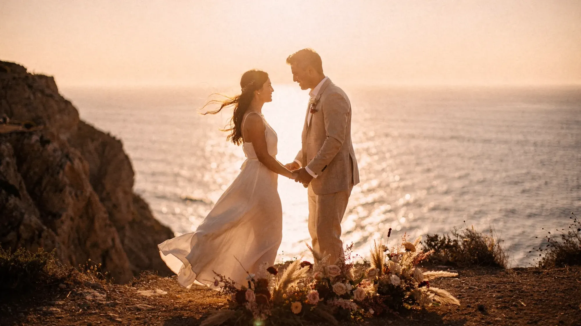 An intimate elopement moment on a Spanish Mediterranean cliffside at golden hour, a couple holding hands with the sea behind them, warm sunlight, windswept hair, minimal florals, cinematic atmosphere.