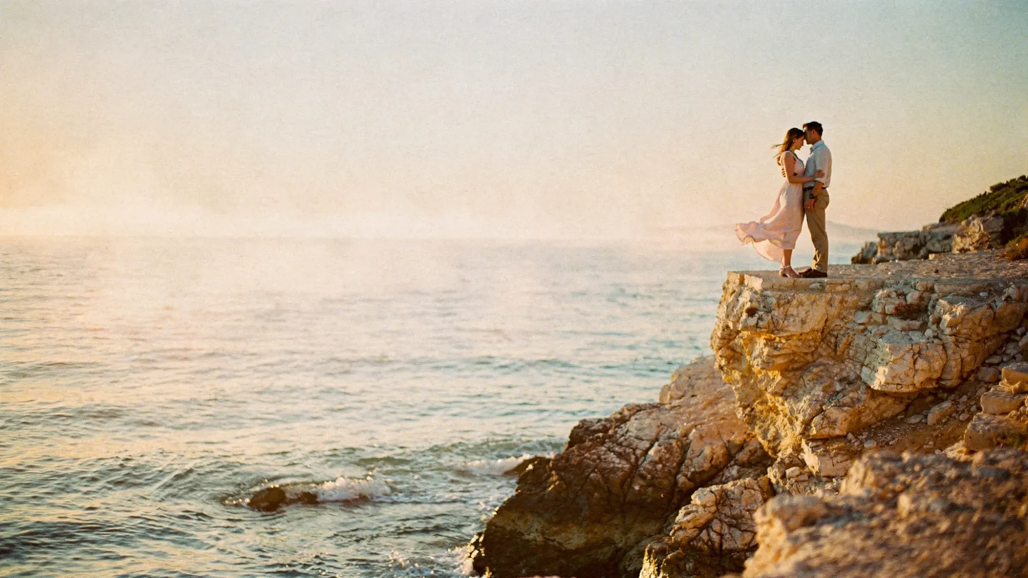 A quiet Mediterranean shoreline at golden hour with soft haze over the sea. A couple stands close together on a rocky overlook above the water, wind moving their clothes slightly. The light is diffused and pastel, with calm reflections on the ocean and a sense of privacy and stillness.