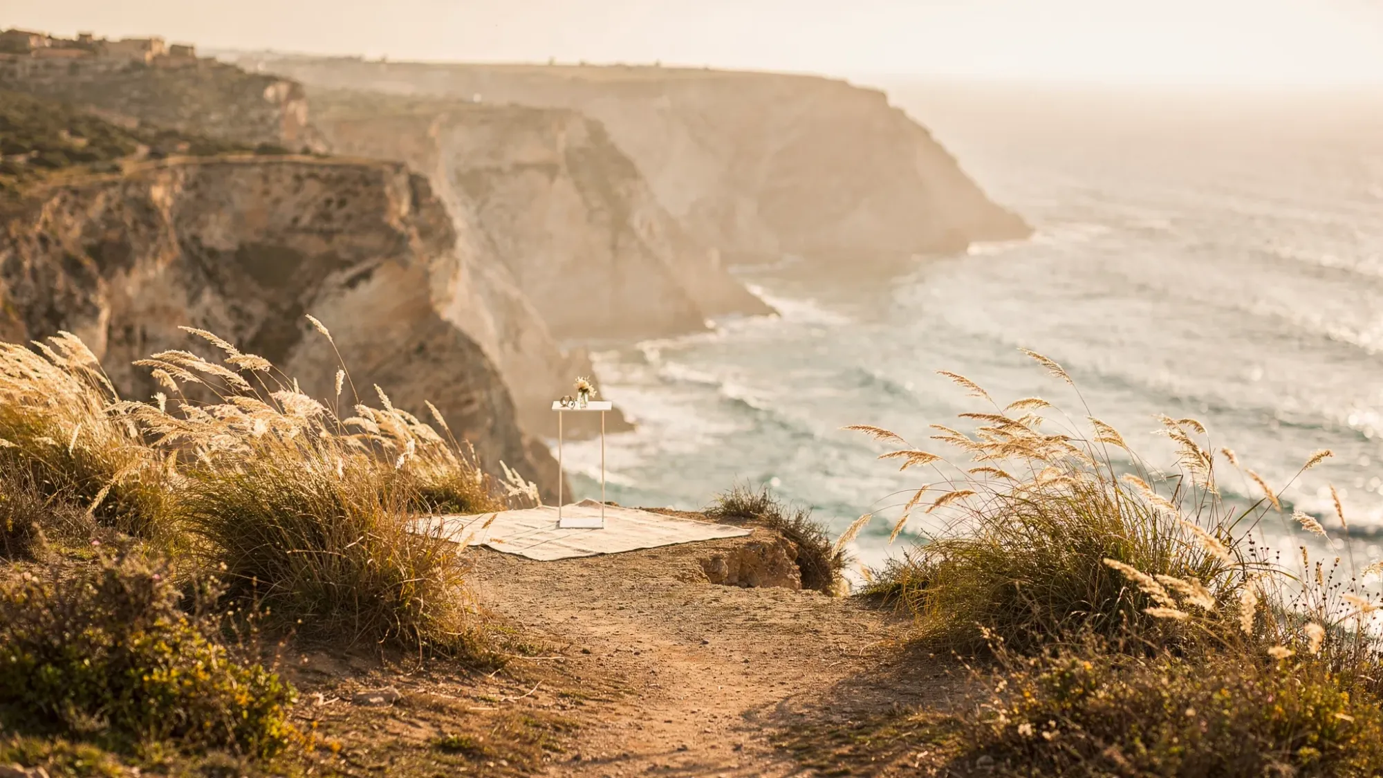 A quiet cliffside overlook on the Spanish Mediterranean coast at golden hour, with a narrow footpath leading to a small flat spot for vows. Wind-bent grasses in the foreground, sea far below, and warm sun spilling across layered cliffs.