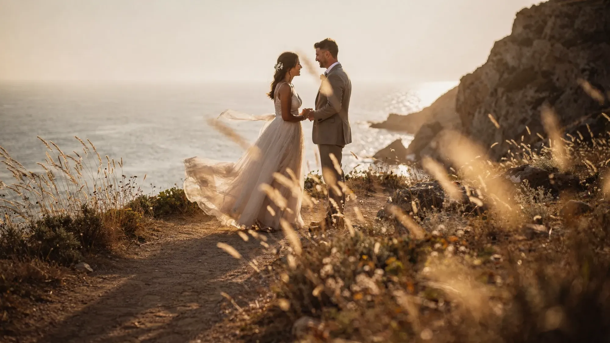 An intimate elopement moment on a Mediterranean cliff above the sea at golden hour, with wind moving through the couple’s clothing, sunlit grasses in the foreground, and a quiet path leading to the vow spot.