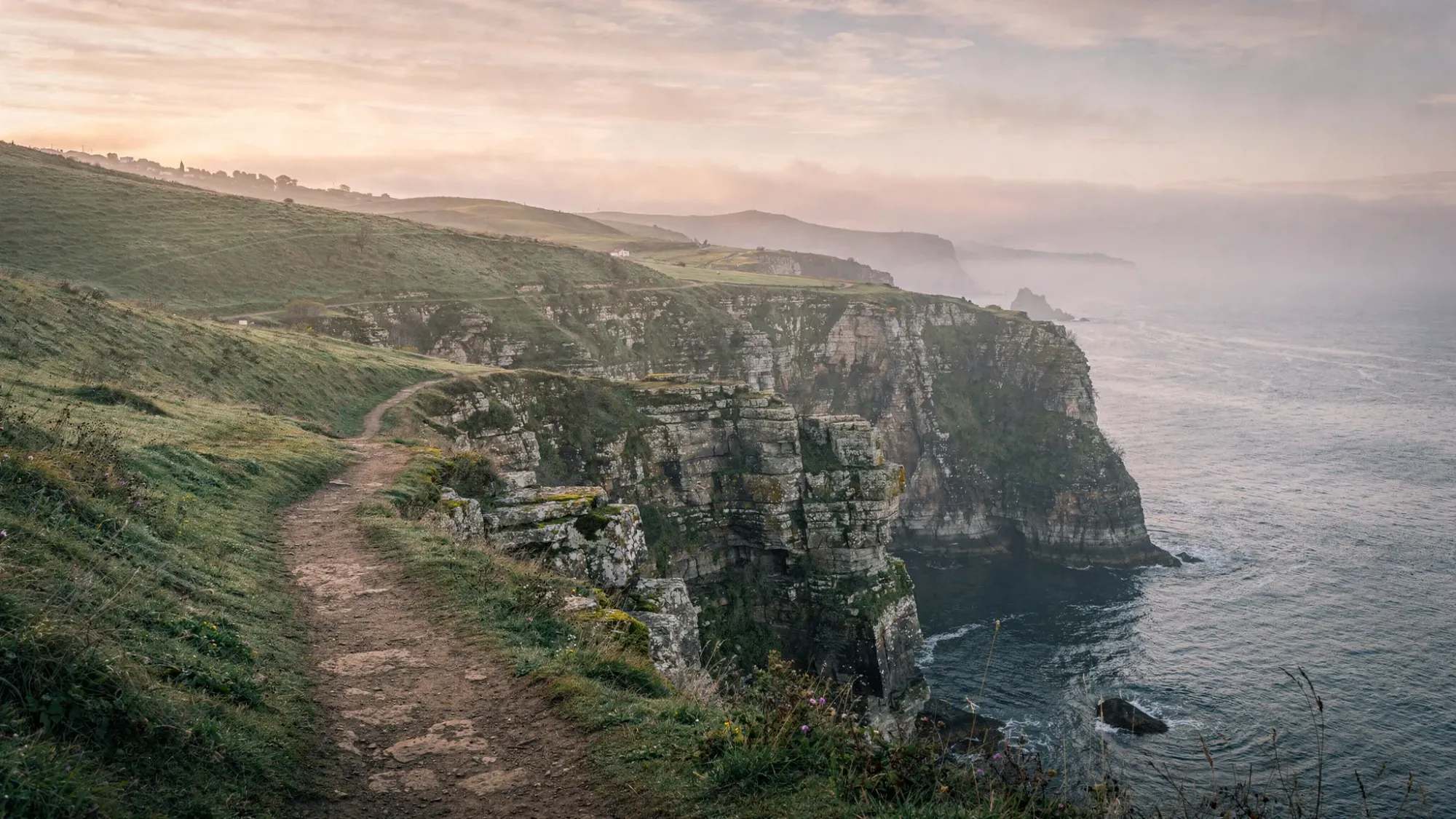 A quiet Atlantic cliffside in northern Spain at dawn, with rolling green hills meeting rugged sea cliffs, soft mist in the distance, and a narrow footpath leading to a hidden cove below.