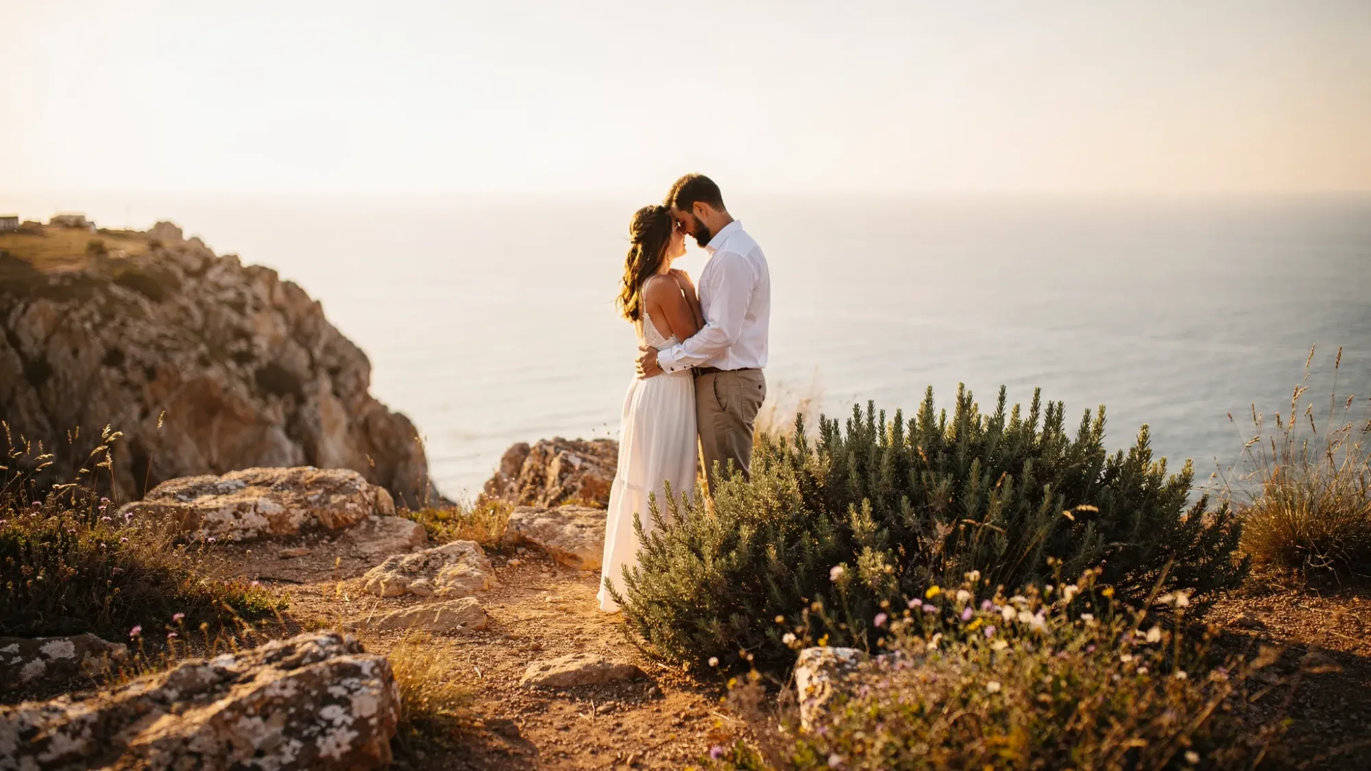 An intimate cliffside elopement moment in coastal Spain at sunrise, with two people standing close near a rugged overlook above the Mediterranean, soft golden light on rock, wild rosemary and grasses nearby, and a calm horizon in the distance.