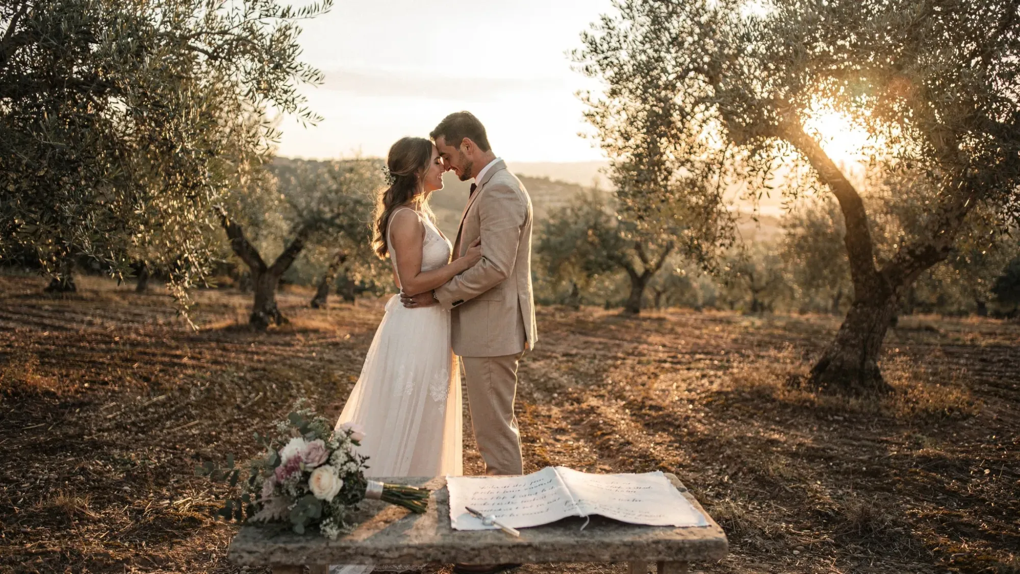 An intimate elopement moment in Spain at sunrise: a couple standing in a quiet olive grove with silver-green leaves, soft golden light, and distant hills, with a minimal ceremony setup like a small bouquet and handwritten vows.