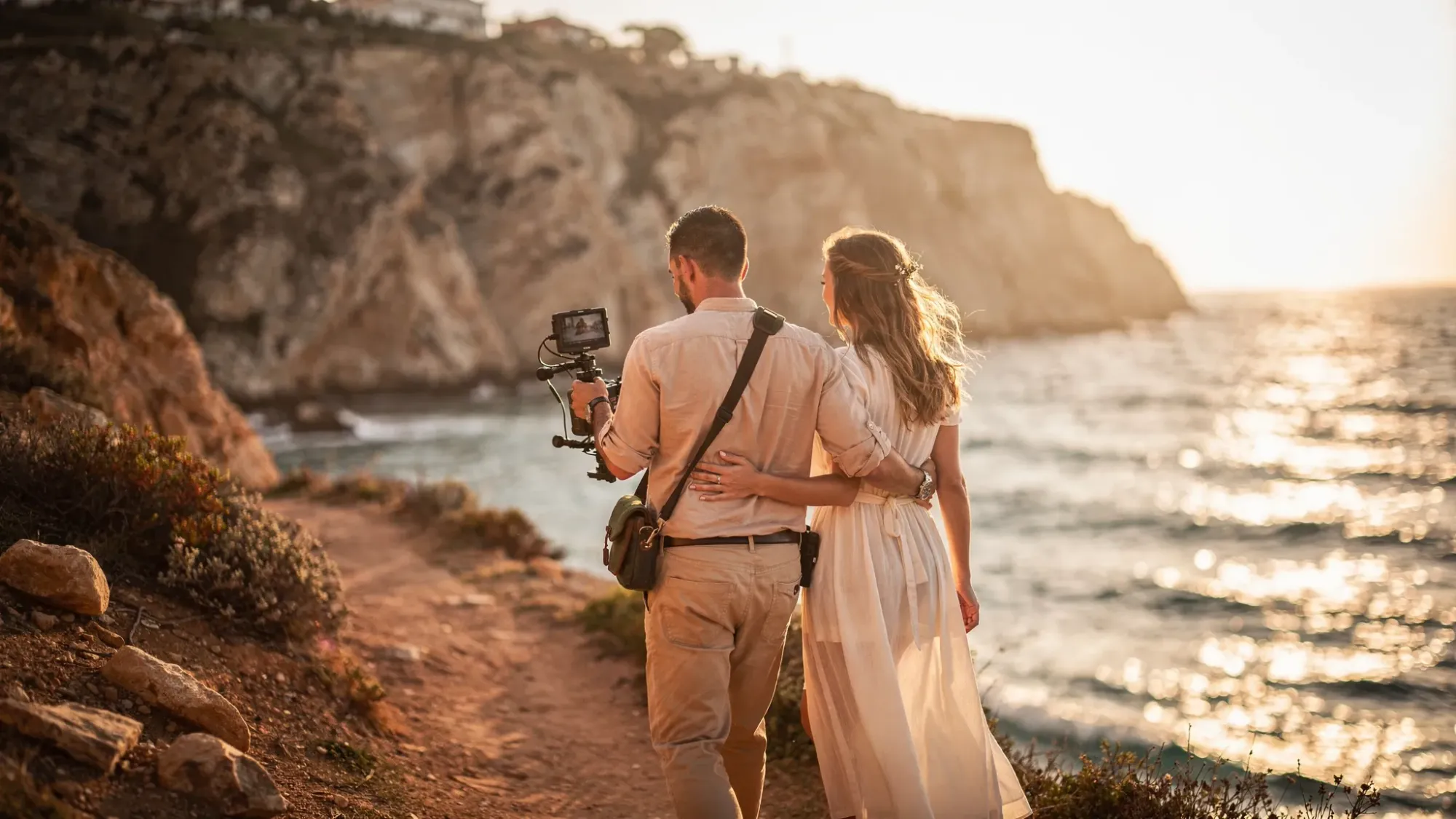 A filmmaker walking with a couple along a narrow coastal path in Spain at golden hour, carrying a compact cinema camera. The couple is relaxed and close, with the sea and rugged cliffs beside them, conveying an intimate, film-first elopement atmosphere.