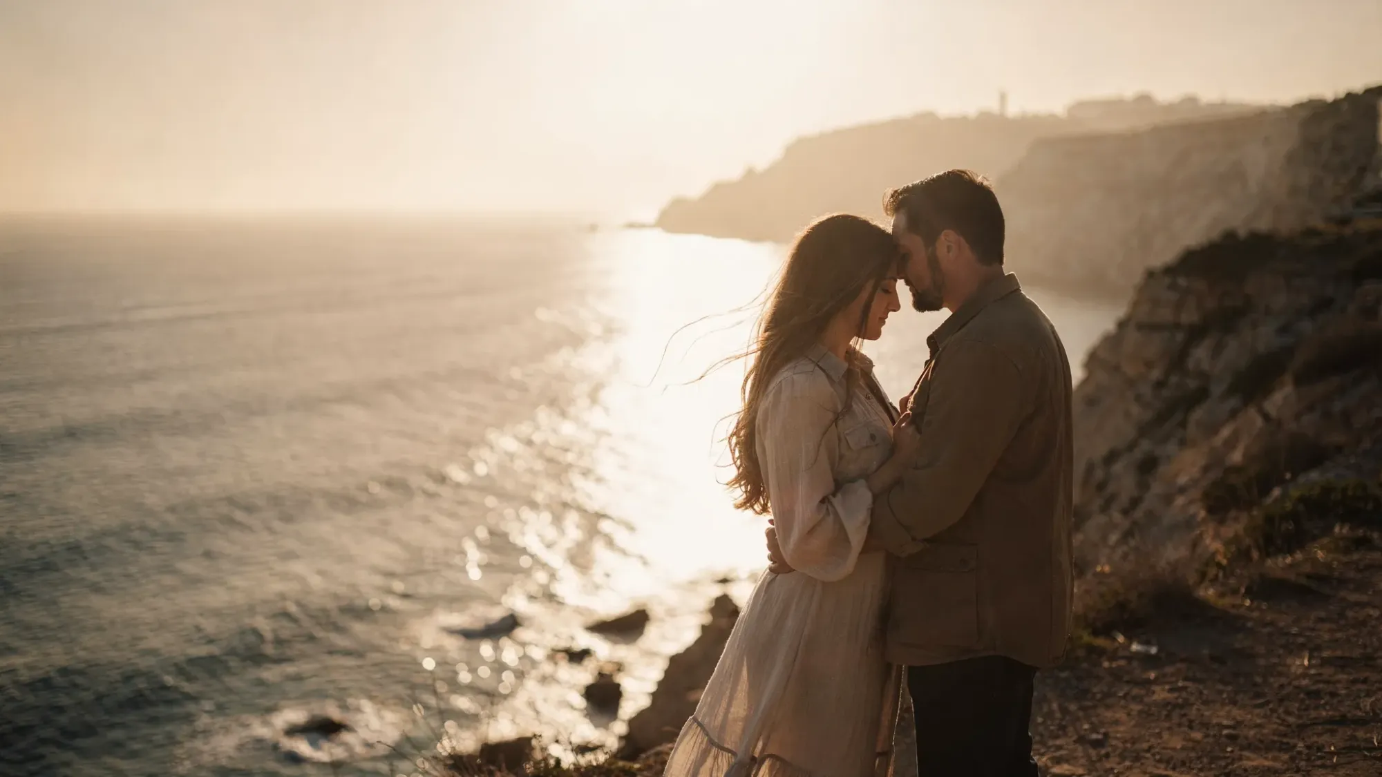 A couple stands close on a quiet Spanish cliffside at golden hour, with wind lifting their clothes slightly. Below them, the Mediterranean glows, and the rocky coastline curves into the distance. The scene feels private and cinematic, with warm light and lots of open space around them.