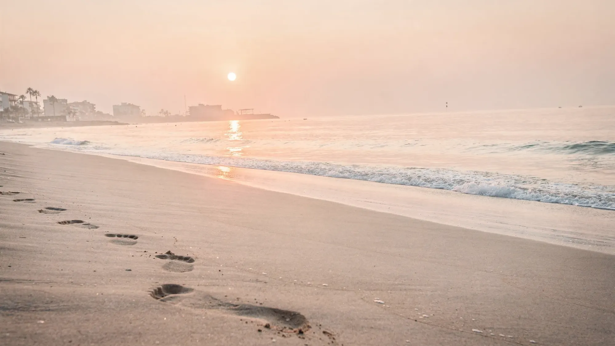 A quiet sunrise on a wide sandy Mediterranean beach in El Campello, with gentle waves, soft peach and gold light on the horizon, and two footprints leading toward the waterline. Far in the distance, low buildings and a few palm silhouettes sit under a pale sky.