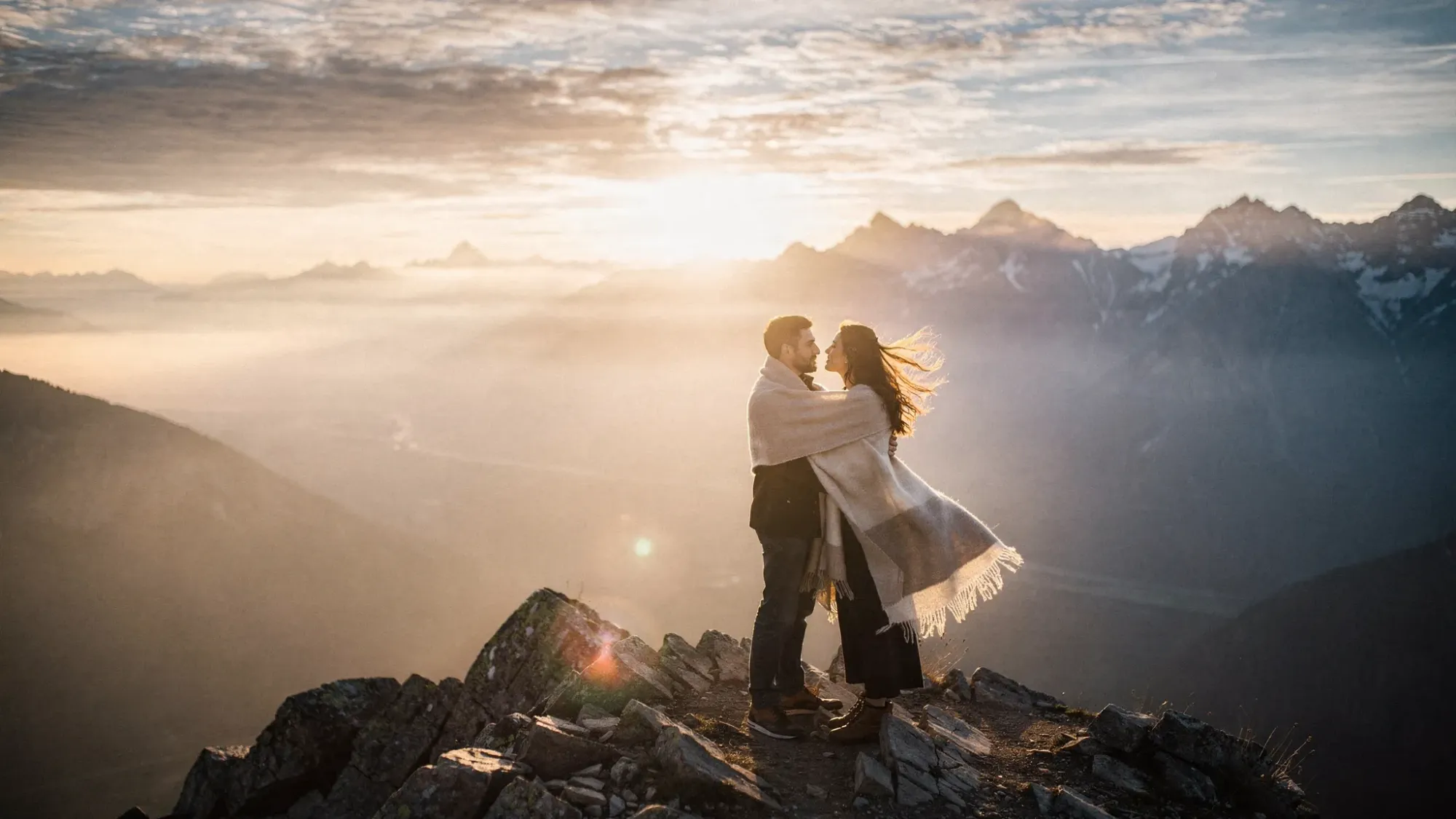 A couple stands on a rocky mountain ridge at sunrise, wrapped in soft layers, with wind moving through their hair. Below them, a wide valley fades into mist, and distant peaks catch the first golden light.
