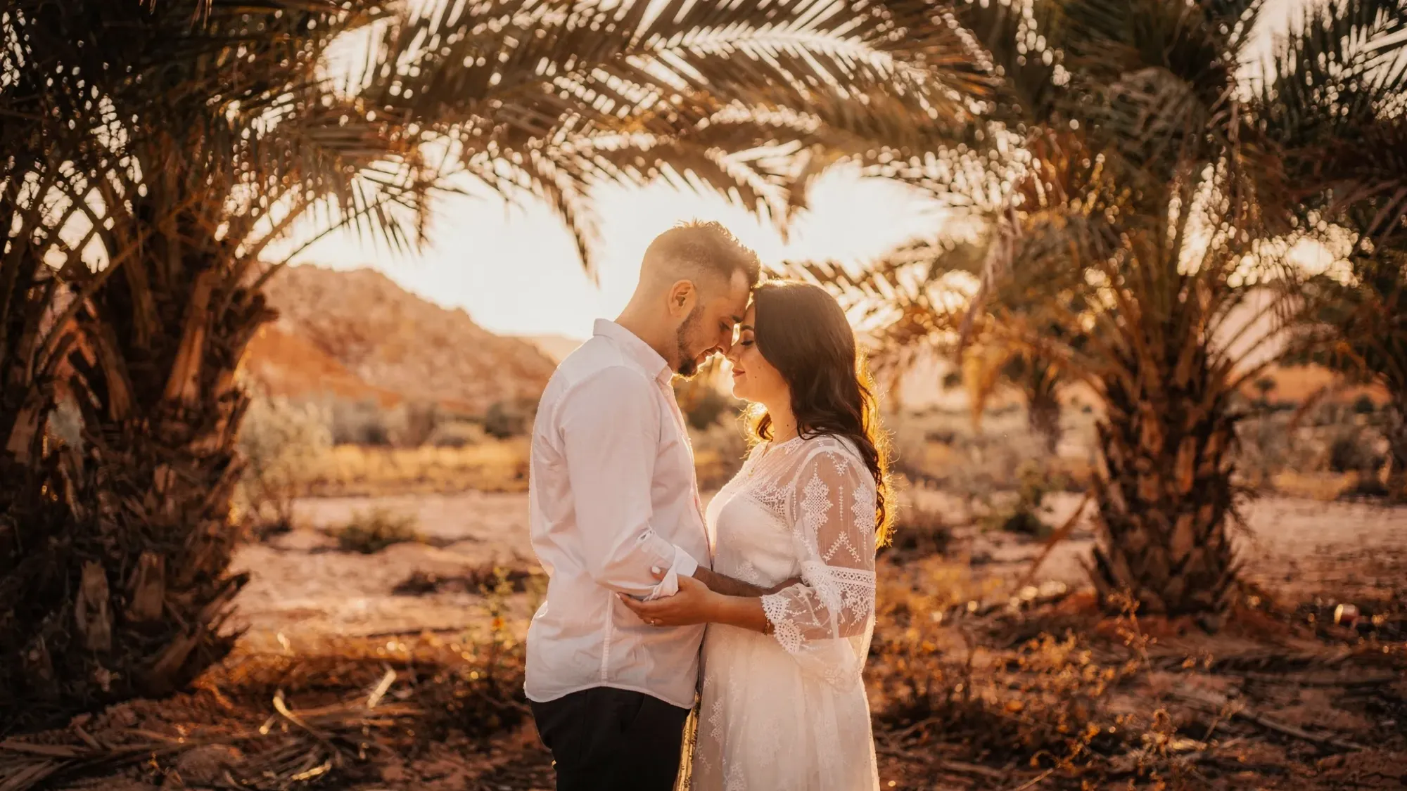 A couple stands close in a hidden palm grove near Crevillente at sunset, hands intertwined, with soft desert light filtering through palm fronds; in the background, warm hills fade into the distance, creating an intimate cinematic frame.