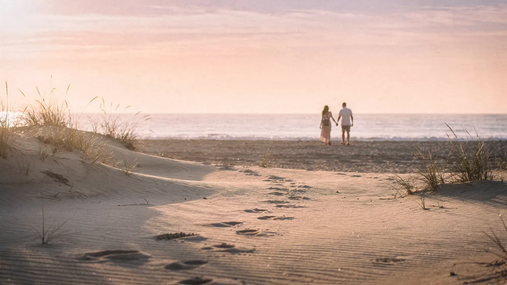 A wide sandy beach with rolling dunes near Gran Alacant at sunrise, soft pastel sky, footprints leading toward the waterline, and a couple walking hand in hand in the distance with the Mediterranean calm and glassy.