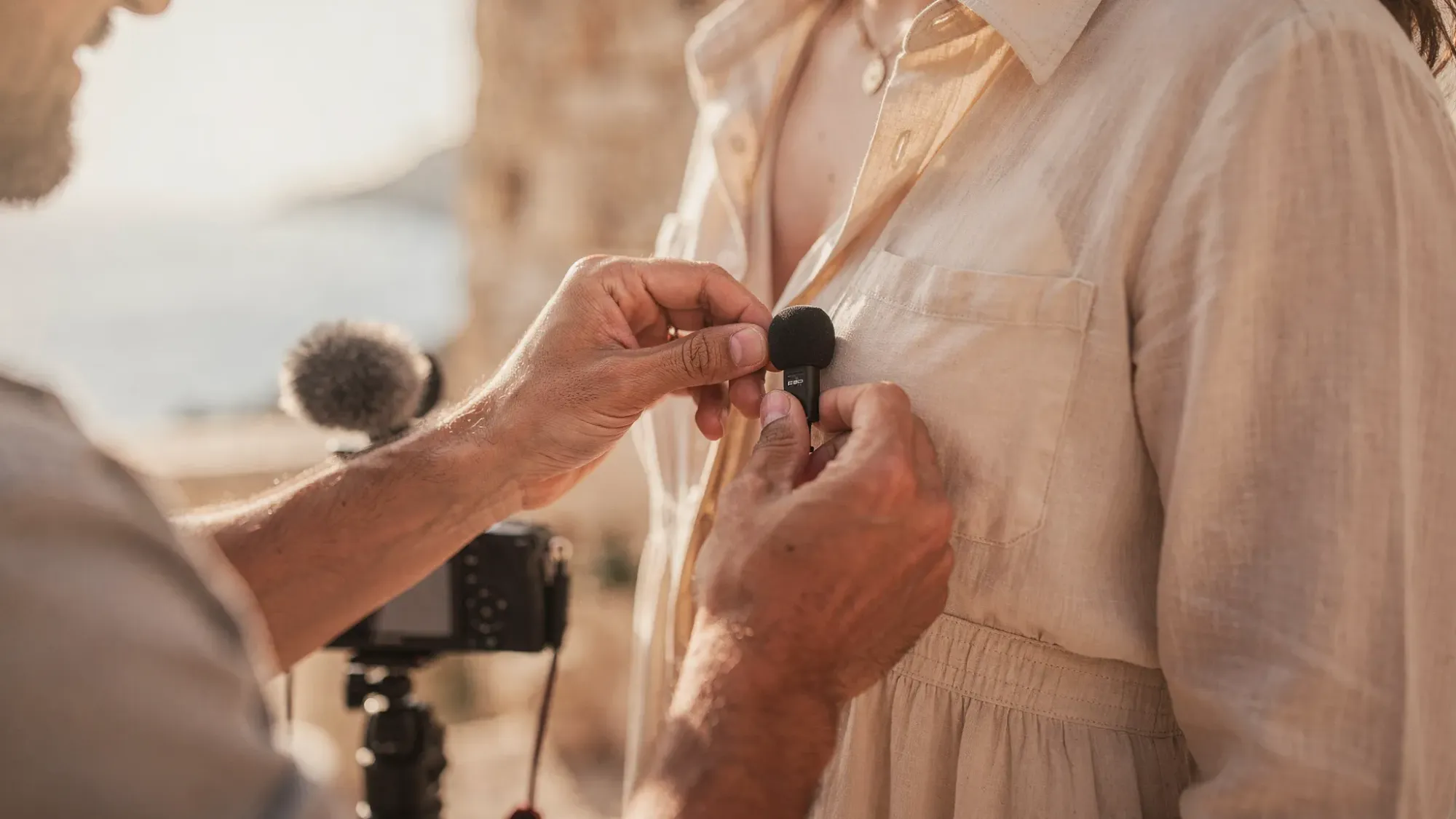 A filmmaker’s hands gently adjusting a small lavalier microphone on a couple’s clothing outdoors, with a compact camera and windscreen visible, and the Mediterranean landscape softly blurred in the background.