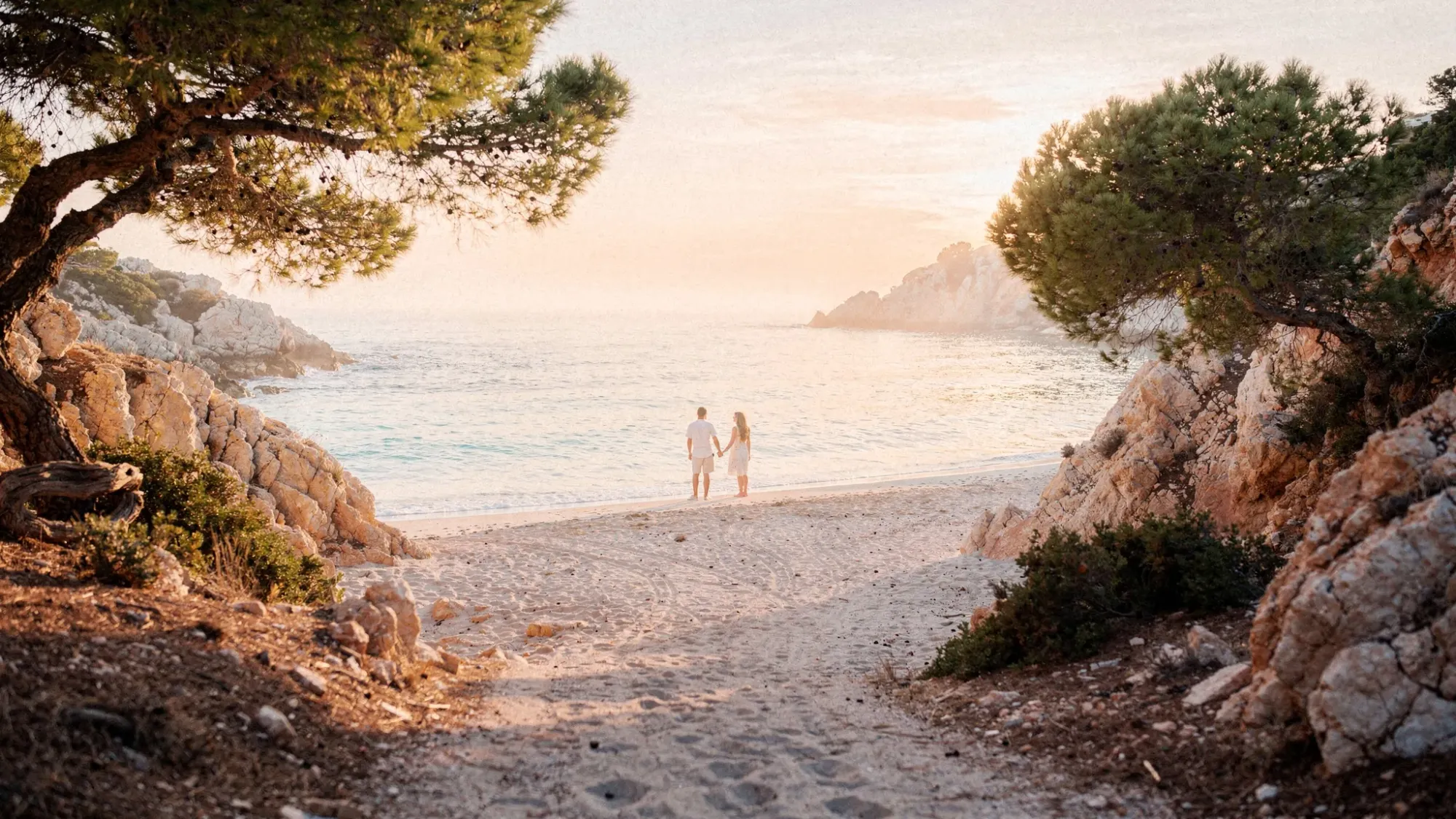 A hidden Mediterranean cove at sunrise with pale sand, calm turquoise water, and a narrow path framed by pine trees and rocky cliffs. Two people stand near the shoreline holding hands, with soft pastel light and a peaceful, empty-beach feeling.