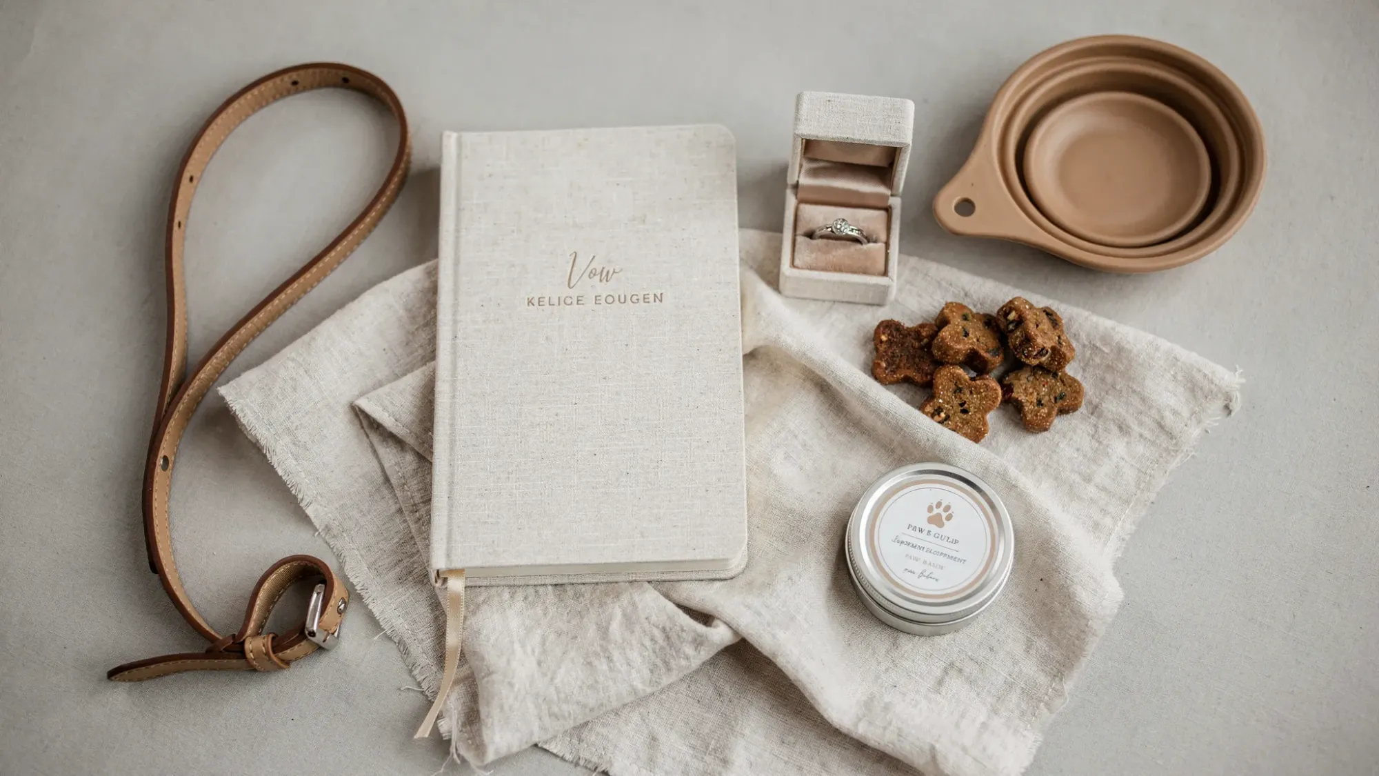 A neatly arranged dog-friendly elopement packing flat lay: vow book, simple rings box, leash, collapsible water bowl, treats, paw balm, and a light linen blanket on a neutral background.
