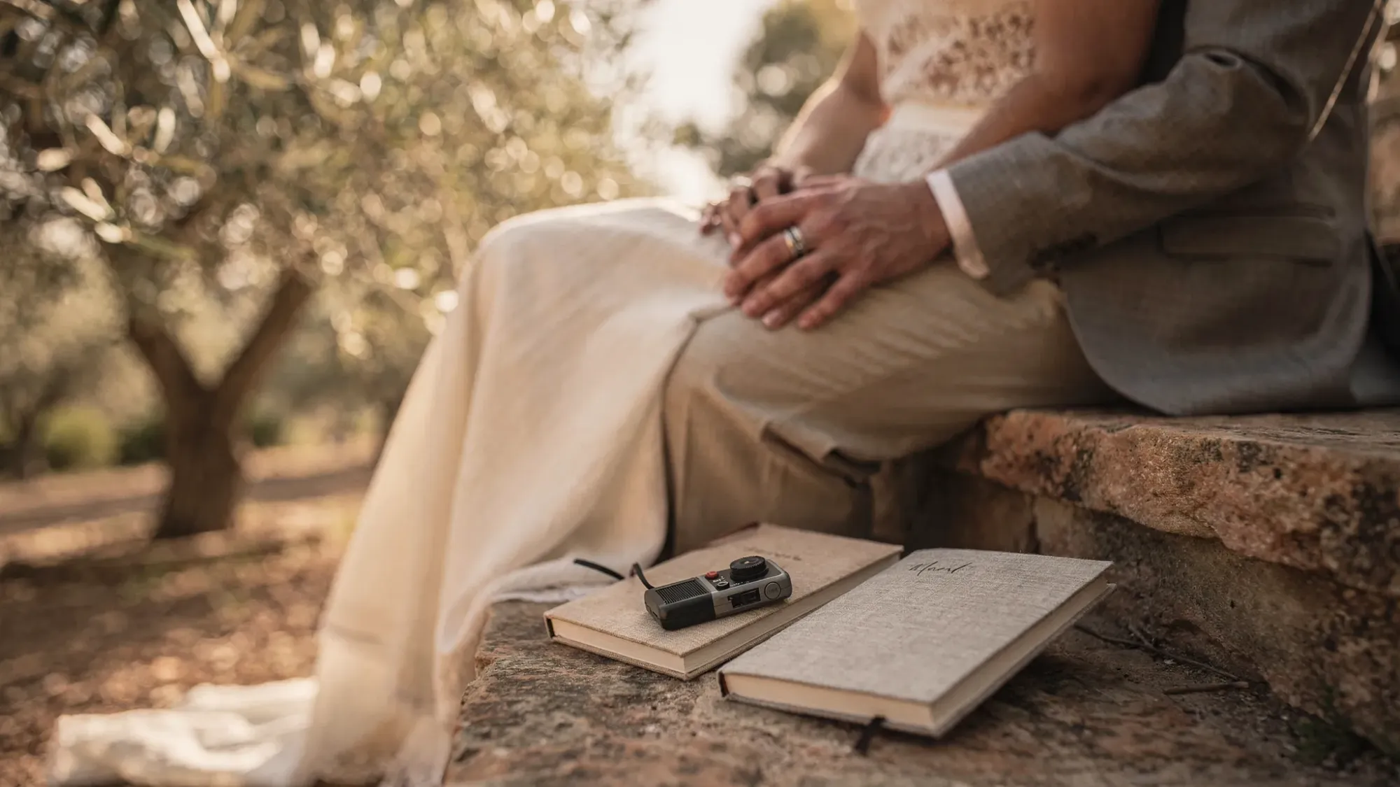 A close, quiet moment after vows with the couple seated on warm stone near an olive grove, hands intertwined, soft late-afternoon light, with a small audio recorder and vow notebooks resting nearby.