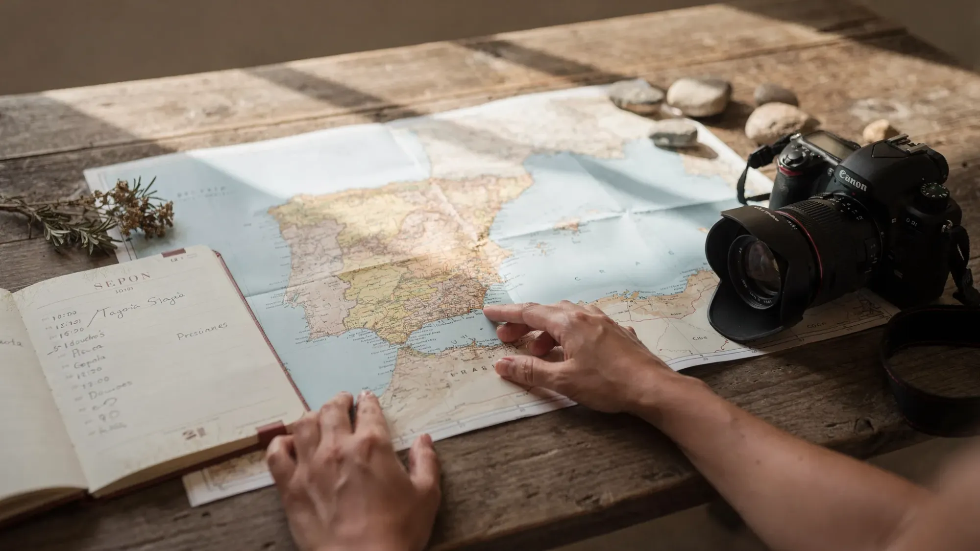 A quiet planning scene for a Spain elopement, with a hands-on map of coastal Spain on a wooden table, a simple timeline notebook, a camera beside it, and small natural details like dried rosemary and sea-worn stones suggesting a cinematic, travel-led elopement plan.