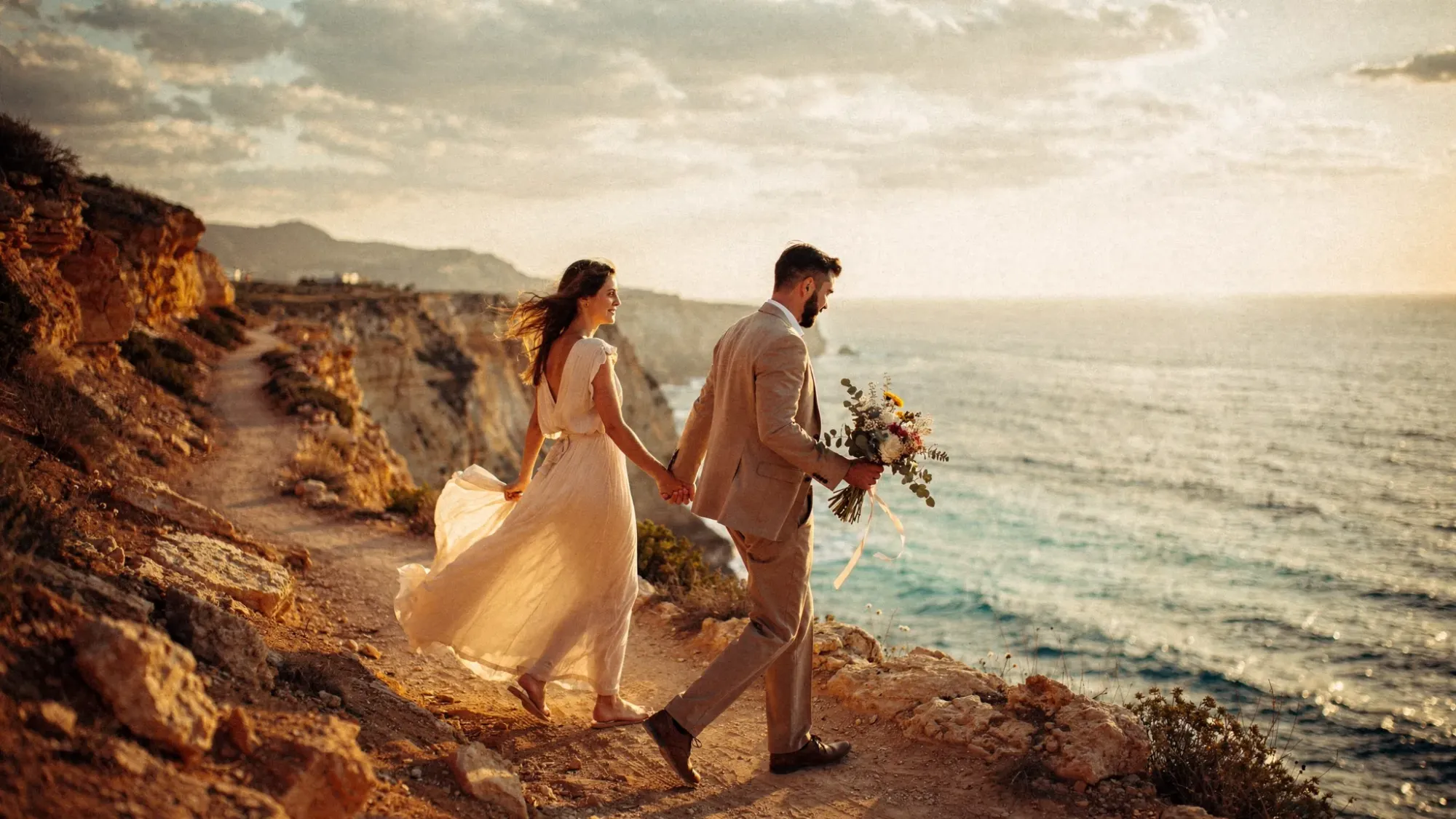 A cinematic Spain coastline elopement scene at golden hour: a couple walking along a rugged cliffside trail above the Mediterranean, warm light on the rocks, wind moving their clothing, with a small bouquet and no crowd in sight.