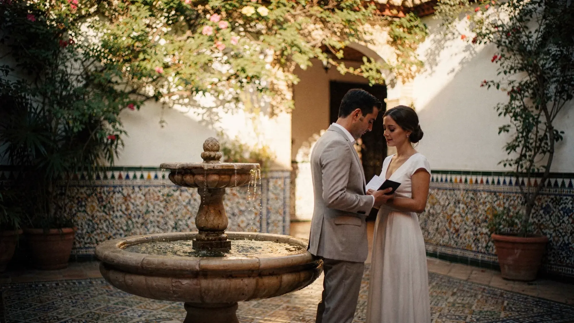 An intimate hidden courtyard in Seville with patterned tiles, a small fountain, and climbing greenery, with a couple standing close together reading vows in soft shade.