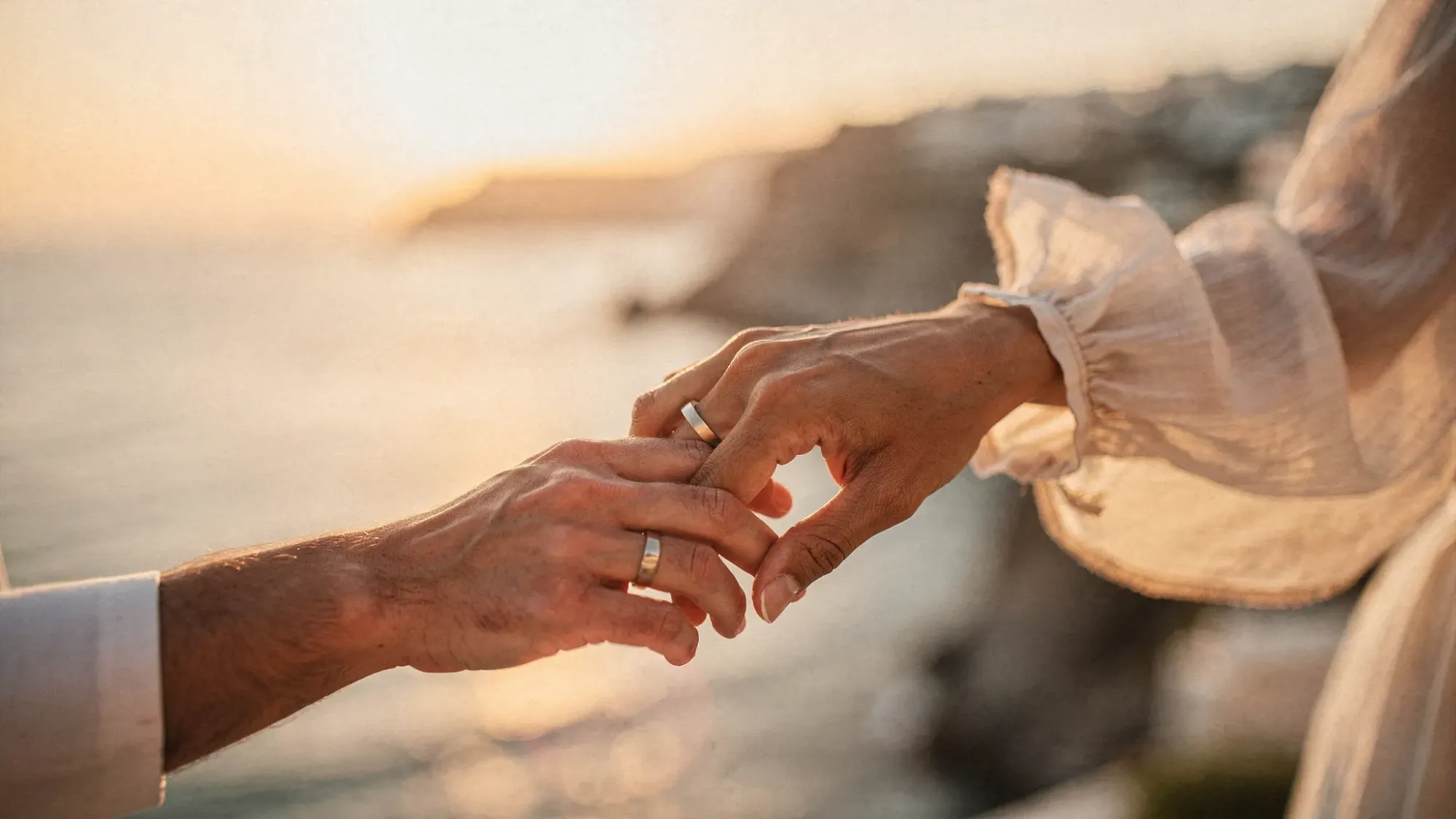 An intimate close-up of two hands holding each other with simple rings, wind moving a sleeve cuff, warm Mediterranean sunset light, and a softly blurred coastline in the background.