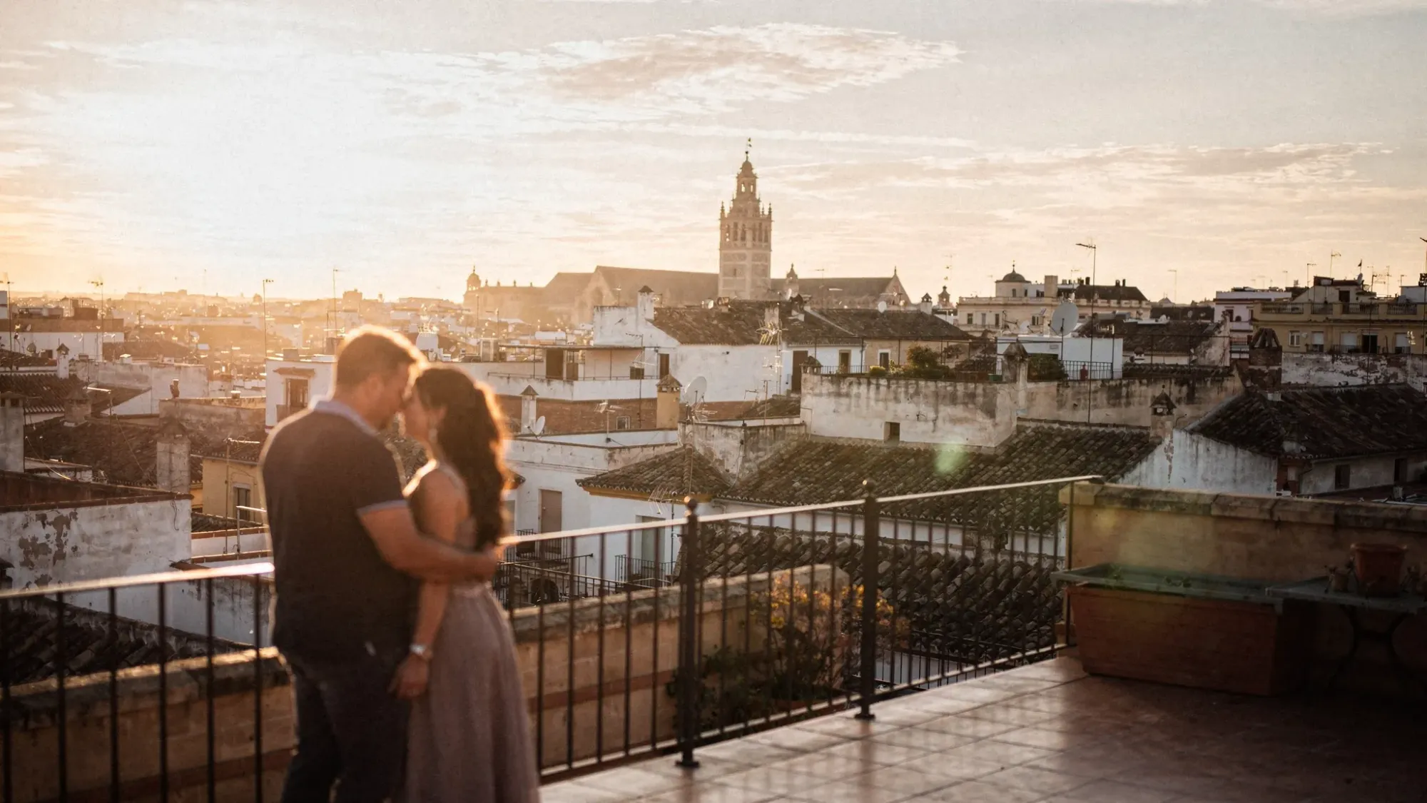 A warm sunset view over Seville’s rooftops with the Giralda tower in the distance, soft golden light in the sky, and a couple leaning on a terrace railing in quiet silhouette.