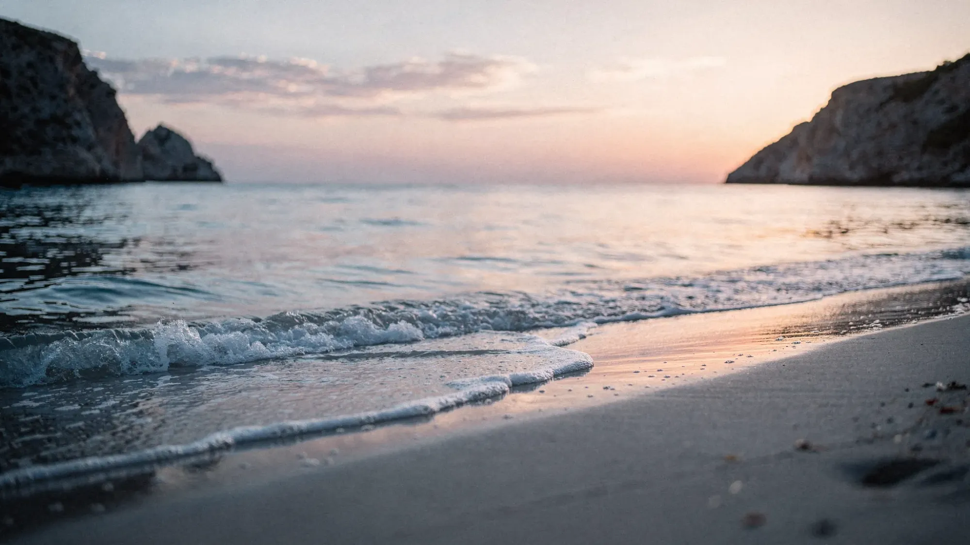 A quiet Mediterranean shoreline at dawn with cool-toned sand, gentle ripples at the waterline, soft pink and blue sunrise sky, and distant cliffs framing a secluded cove. No crowds, no chairs, just open space and calm light.