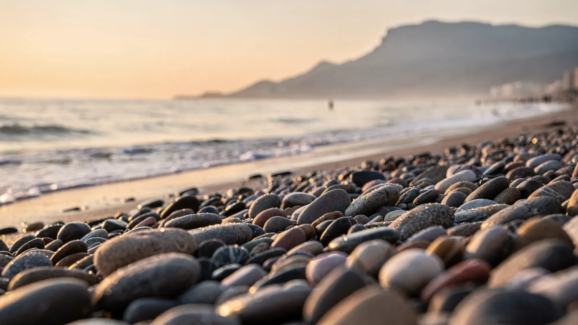 A quiet morning on Playa del Albir with smooth pebbles in the foreground, a calm Mediterranean sea, soft sunrise light, and a mostly empty shoreline with distant mountains fading into haze.