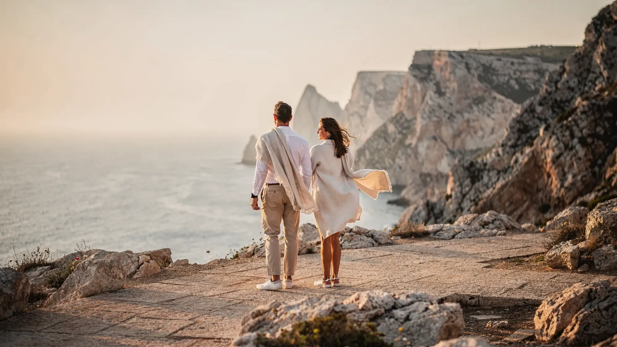 A couple standing on a wide limestone cliff path in Sierra Helada overlooking the Mediterranean, wind moving their clothes and hair, with rugged coastal cliffs and calm blue sea below in early morning light.