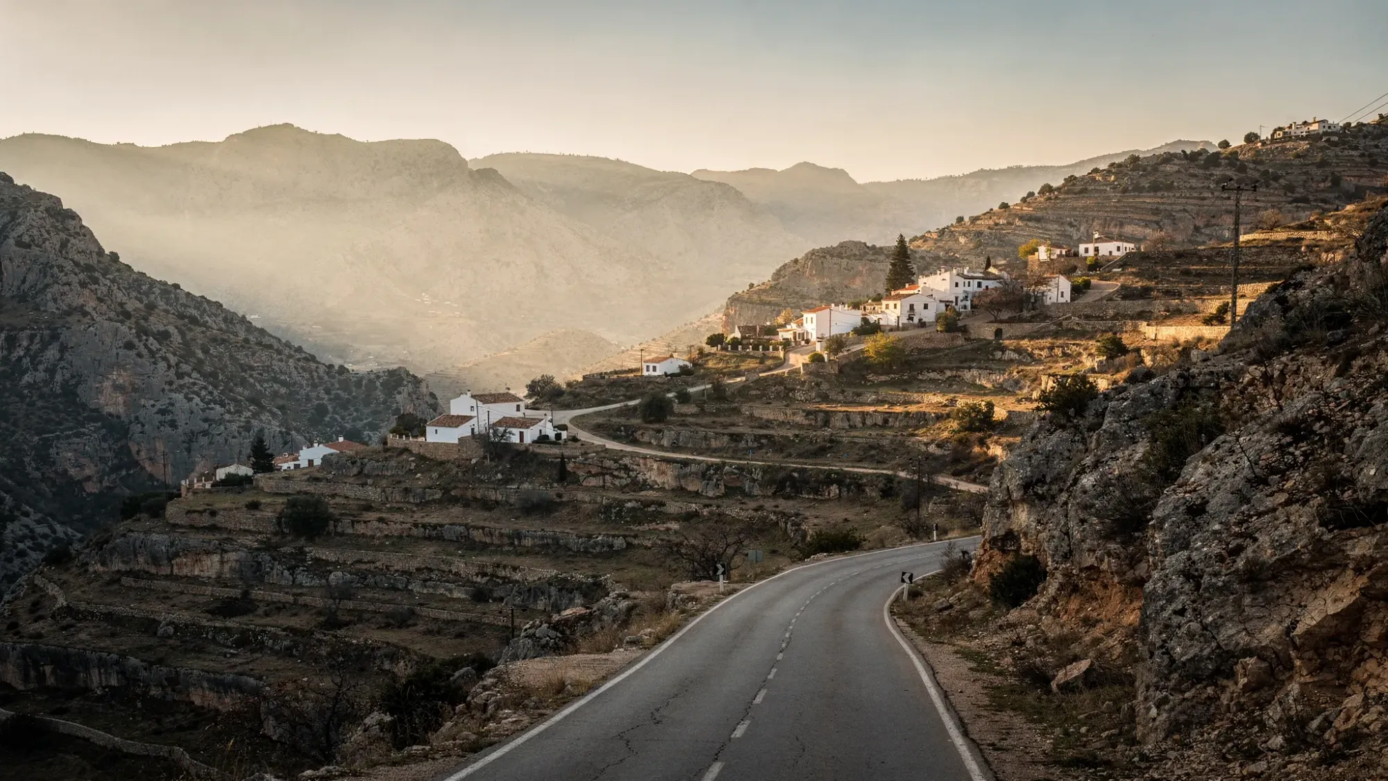 A quiet mountain valley in Vall de Laguar at golden hour, with layered limestone ridges, terraced hillsides, and a narrow winding road leading toward small white villages perched on the slopes. The light is soft and warm, with long shadows and a calm, intimate atmosphere.