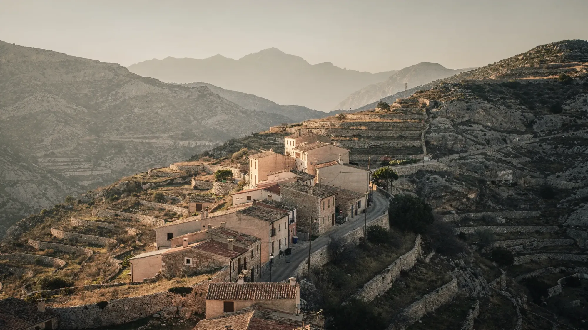A quiet mountain village perched on a ridge in Alicante province, Spain, with stone houses and terraced hills falling away into a wide valley. Warm golden light touches the rooftops, and distant mountains layer into a hazy horizon.