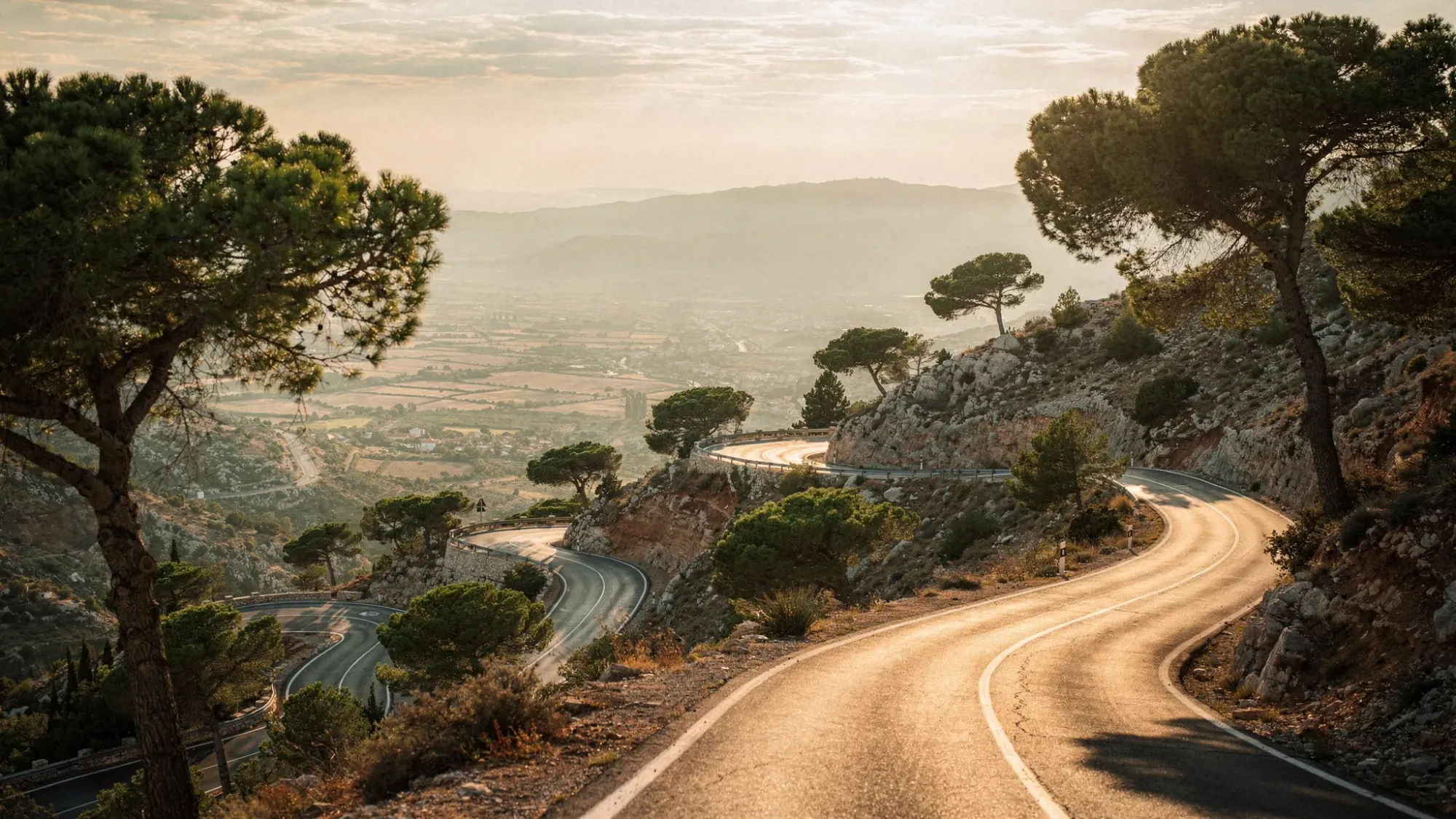 A winding mountain road with dramatic hairpin turns climbing through Mediterranean pines, with a wide valley view opening in the distance under soft golden-hour light.