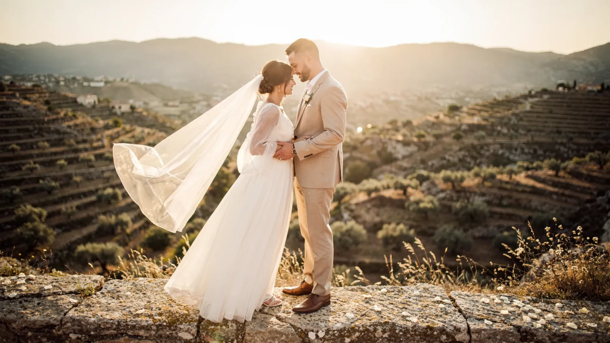 An intimate elopement couple stands on a quiet limestone overlook above a terraced Mediterranean valley, with almond and olive groves below and distant mountains fading into warm golden-hour light. The scene feels private and cinematic, with wind gently moving fabric and no crowds in sight.