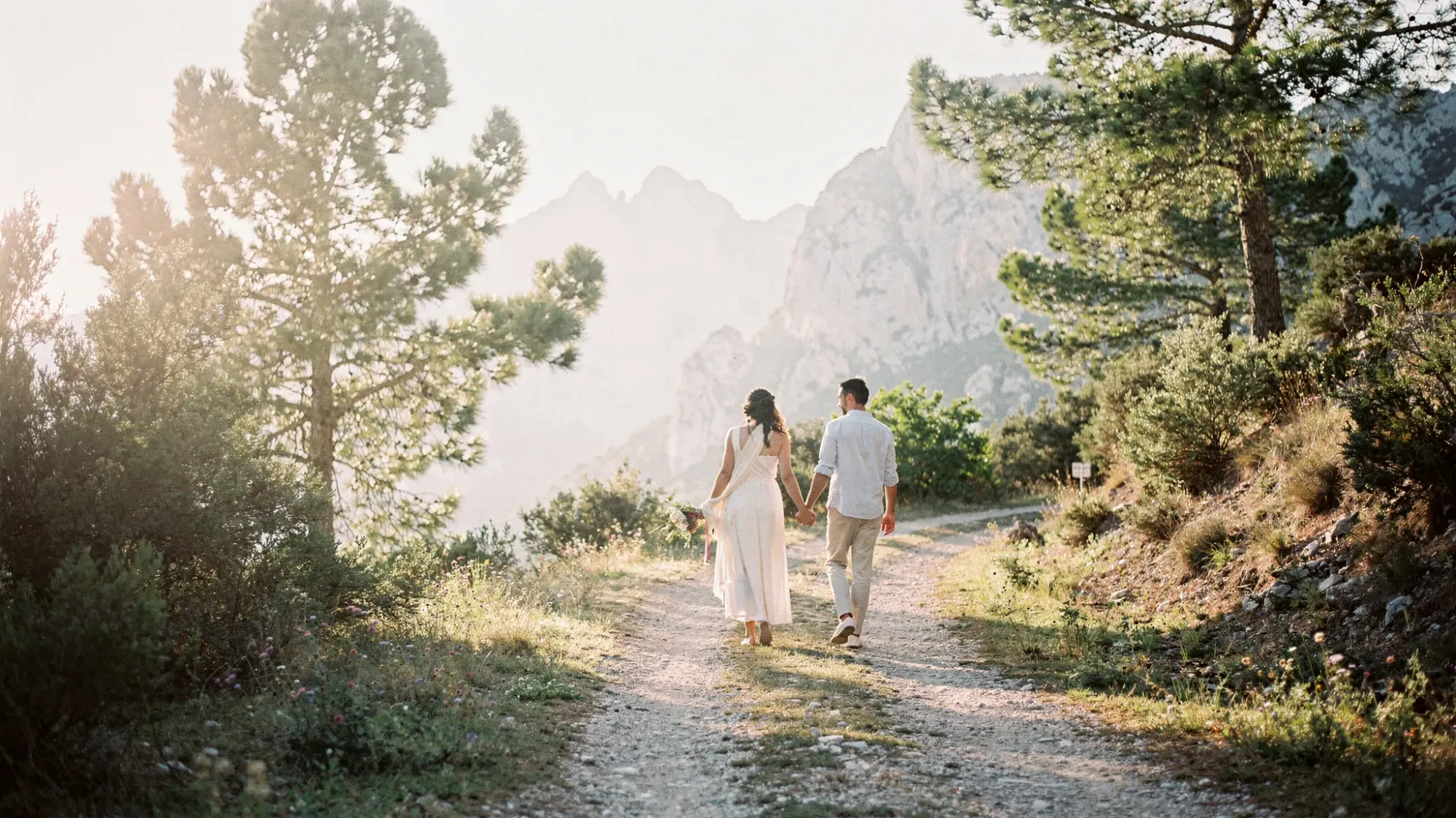 A quiet mountain trail near a tiny Spanish village, lined with pine trees and low shrubs, opening toward pale limestone peaks. A couple in simple elopement attire walks hand in hand in soft sunrise light, with no crowds or buildings in sight.