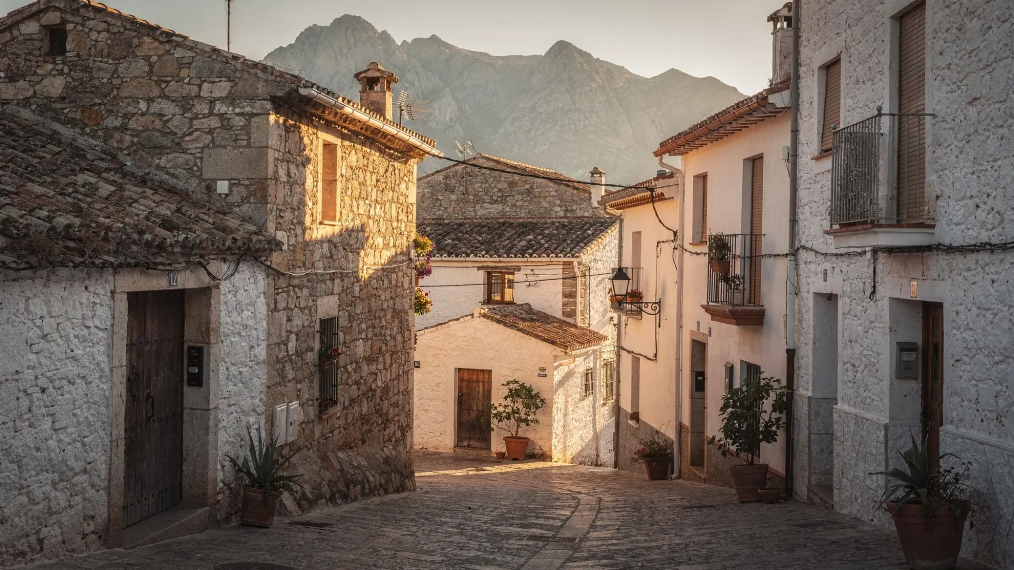 A quiet stone mountain hamlet in southern Spain at golden hour, narrow lane with rustic houses, terracotta roofs, distant rugged peaks, warm sunlight pooling between buildings, no people visible.