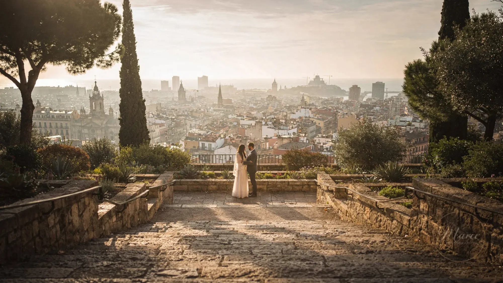 A quiet sunrise scene in Barcelona on Montjuïc overlooking the city and the sea, with warm golden light spilling across stone steps and gardens. A couple stands close together in the distance, small in the frame, surrounded by soft shadows and early morning stillness.