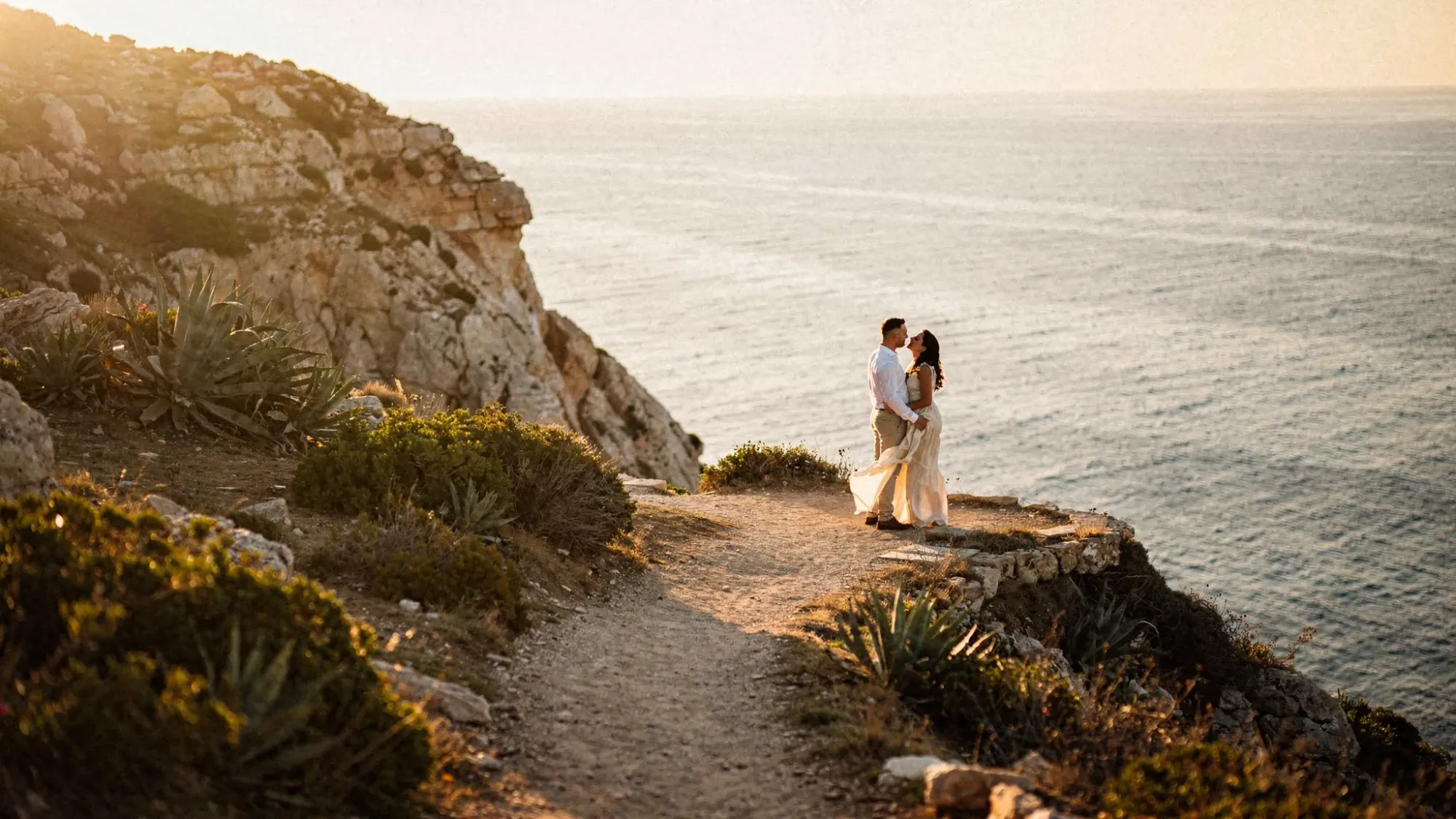A quiet Mediterranean coastal viewpoint at golden hour with a narrow path through low shrubs, warm sun on rocky cliffs, and a calm sea in the distance. A couple stands close together near the edge, small in the landscape, with wind moving gently through their clothing.