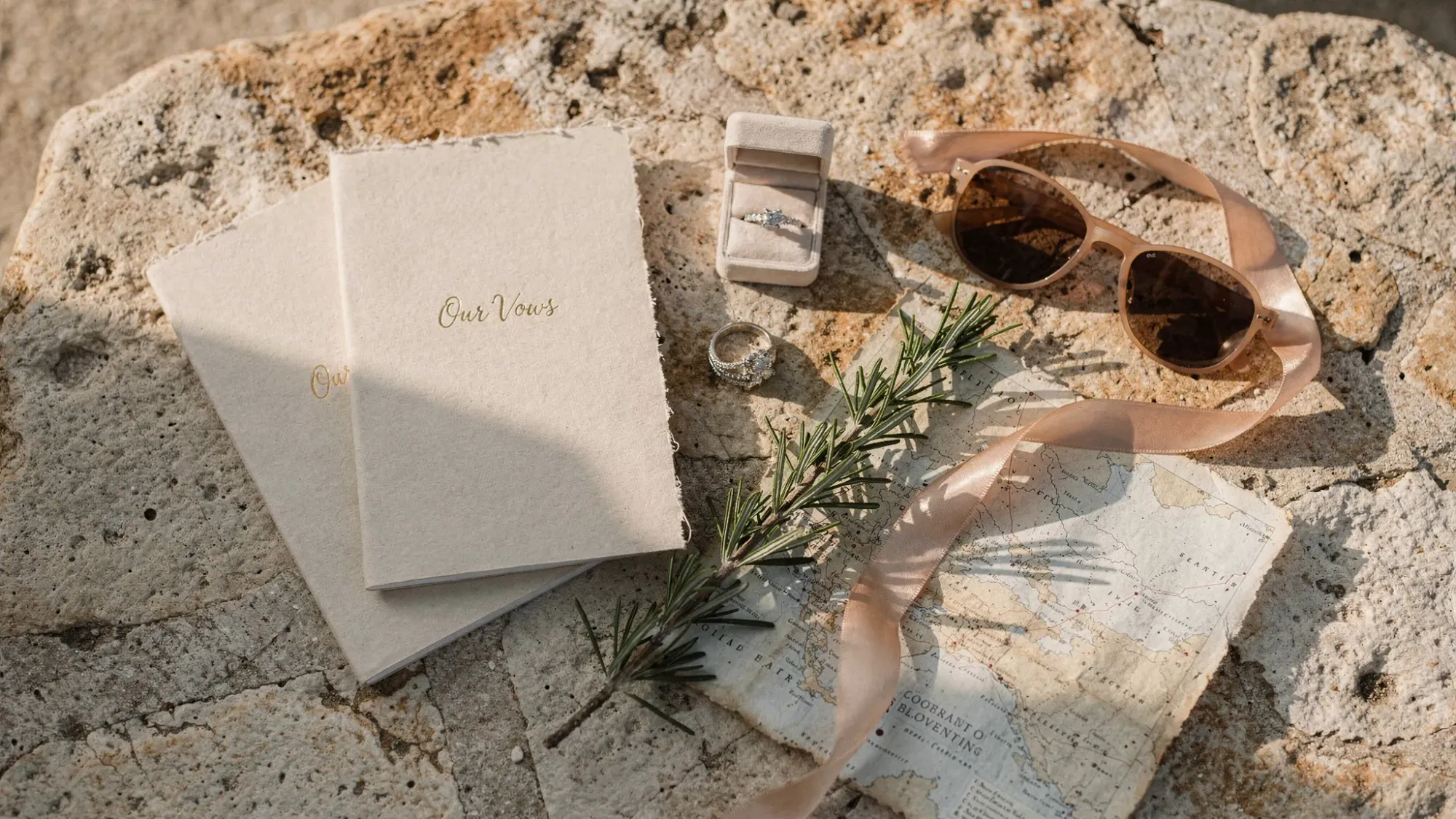 A flat lay of elopement details on Mediterranean stone: two vow booklets, rings, a sprig of rosemary, a small ribbon, sunglasses, and a folded map. Warm natural light falls across the textures, evoking a calm, intimate planning mood.