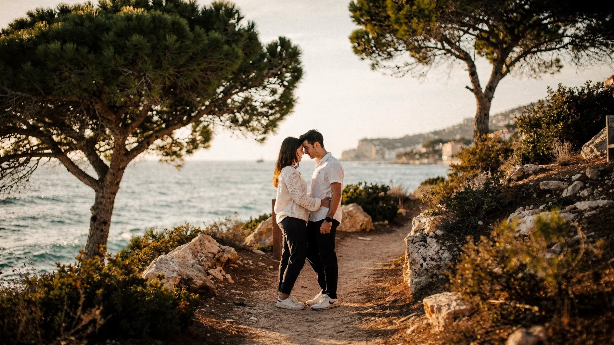 A couple standing close on a coastal cliff path near Albir with pine trees nearby, the Mediterranean stretching behind them, late afternoon soft light, and an empty trail that feels secluded.