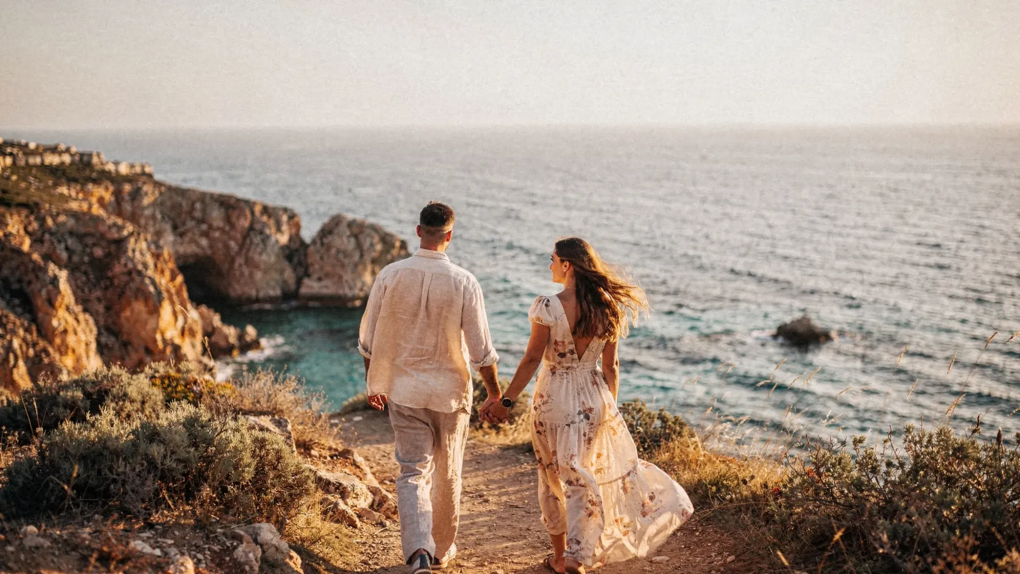 A couple walking hand-in-hand along a coastal path above a Mediterranean cove during golden hour, with warm light on the cliffs, wind moving through their clothing, and the sea stretching into the distance.
