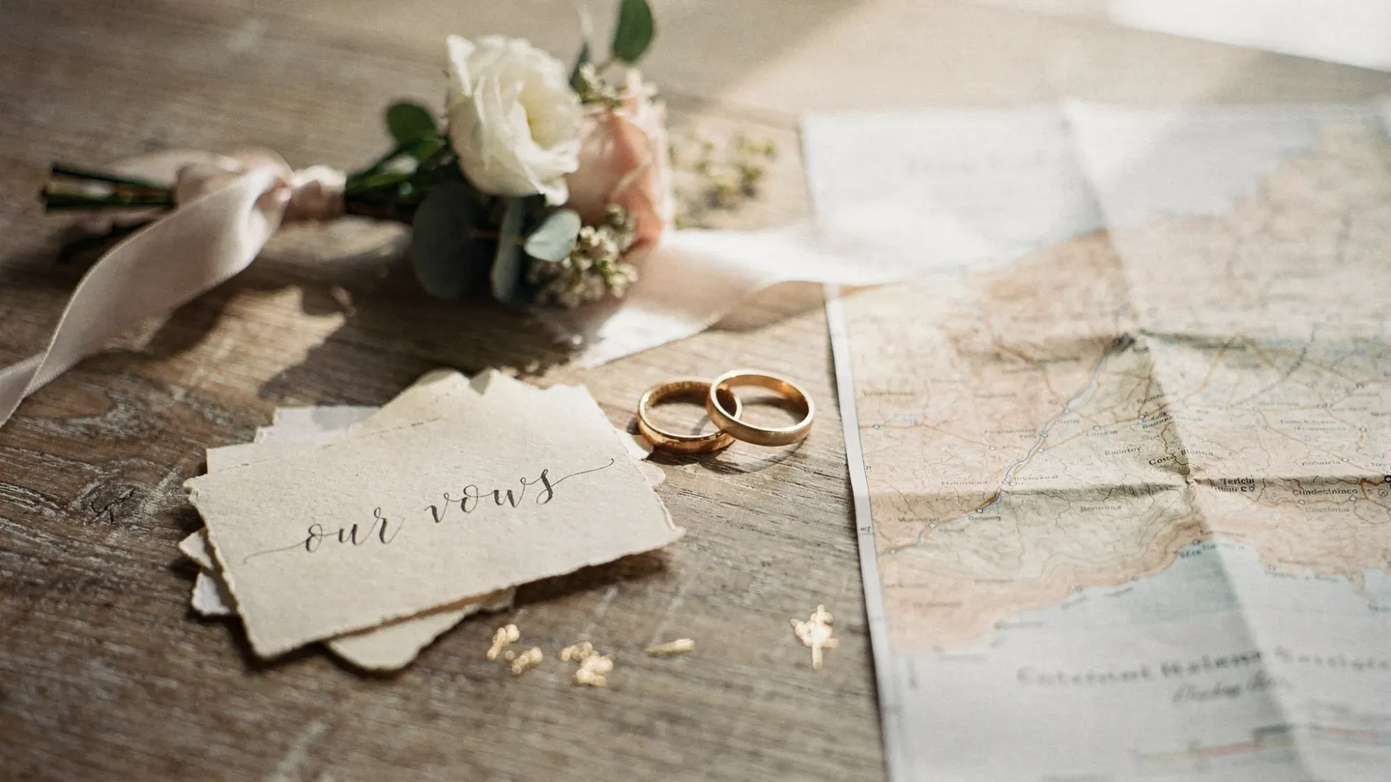 A minimalist elopement timeline concept: two rings, handwritten vows, a small bouquet, and a map of inland Costa Blanca on a rustic table with soft natural light.