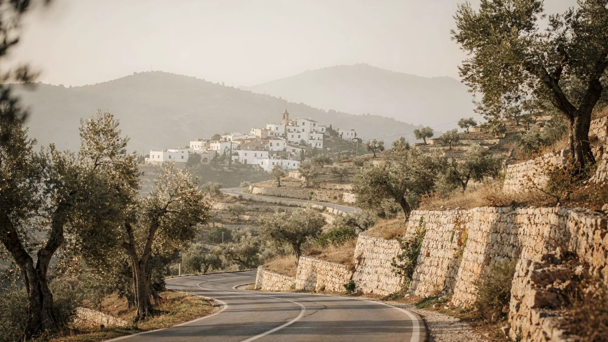 A narrow mountain road winding through a Mediterranean valley with stone terraces, small white village rooftops in the distance, olive trees along the roadside, and soft morning mist in the hills.