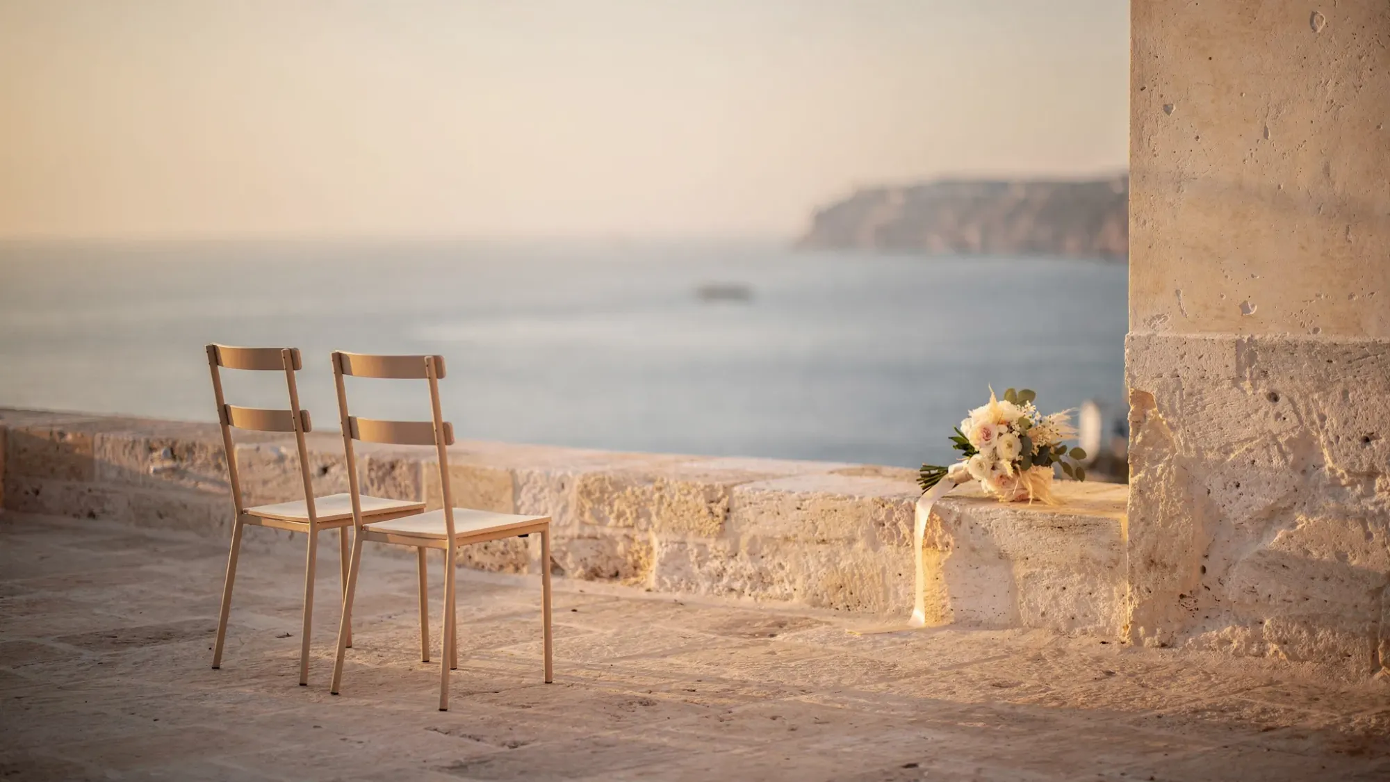 A quiet Mediterranean vow setup on a secluded stone terrace overlooking the sea at sunset, with two simple chairs, a small bouquet on a stone ledge, and soft golden light hitting textured walls; no crowd, no decorations beyond a few intentional details, emphasizing intimacy and space.