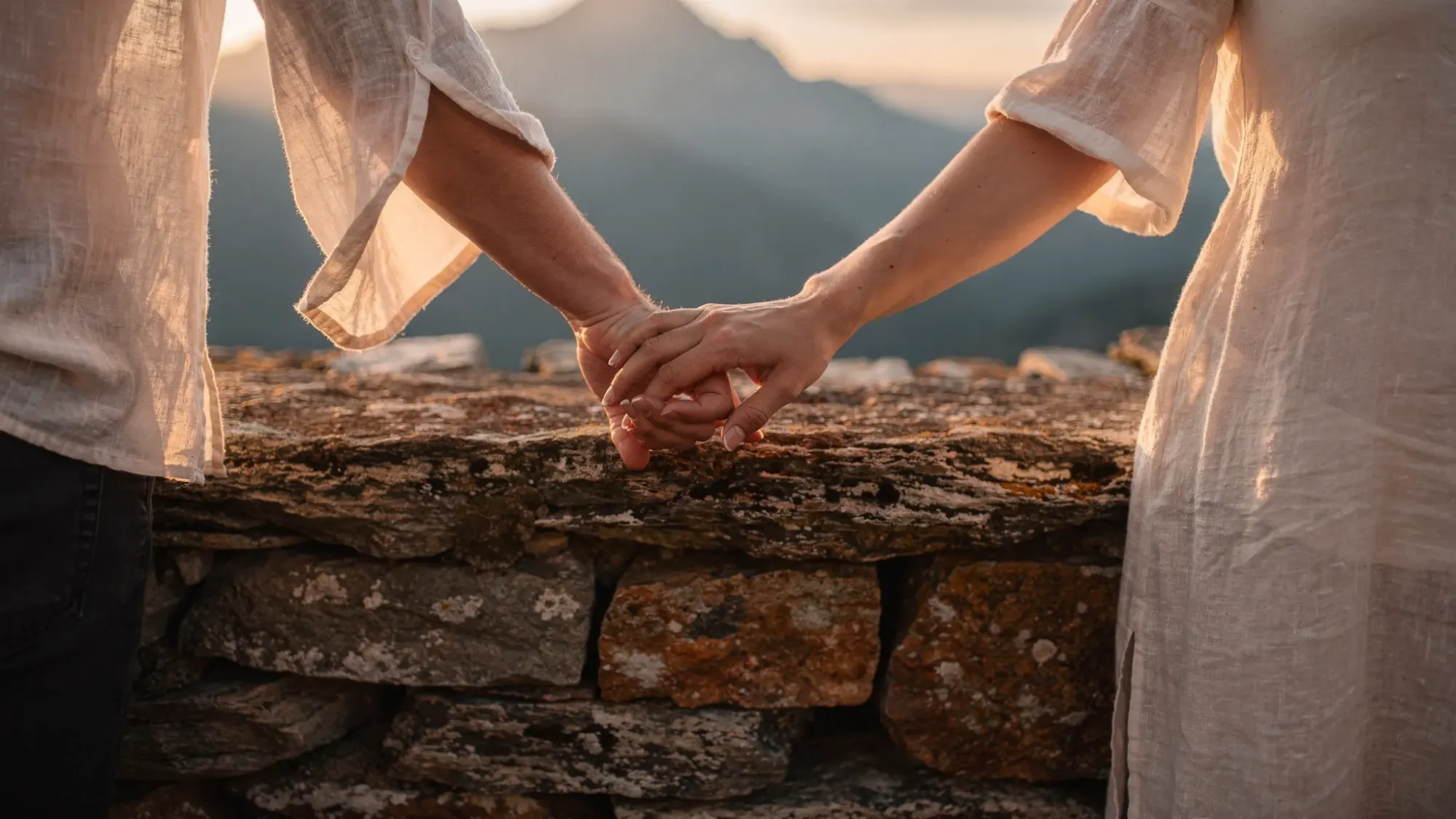 A close, intimate moment of a couple holding hands on a stone terrace overlooking a mountain valley, with wind gently moving their clothing and soft evening light catching the texture of the dry-stone wall.