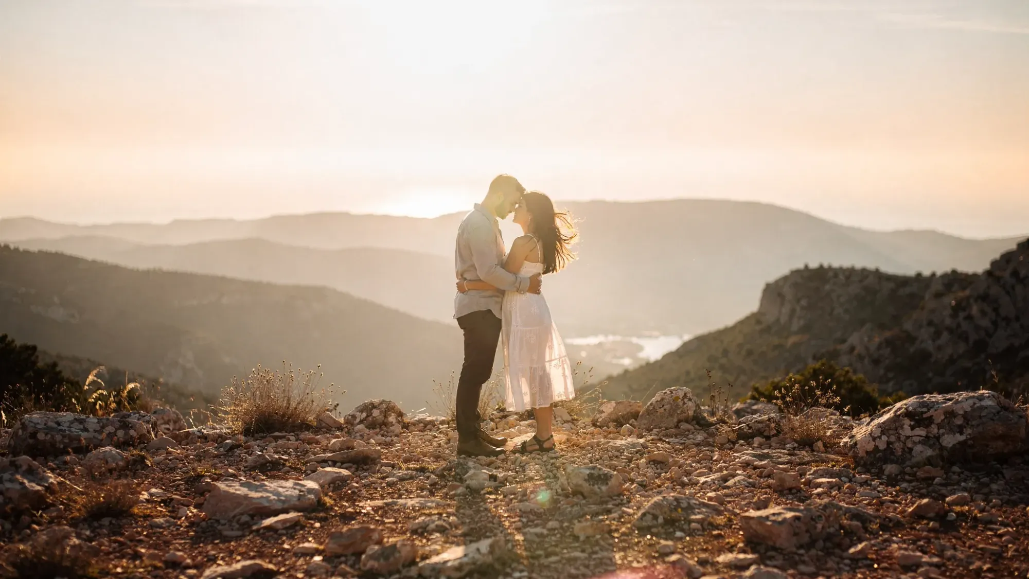 A quiet mountain overlook near Tàrbena at golden hour, with rolling hills fading into the distance, warm light on rocky ground, and a couple standing close in the wind.