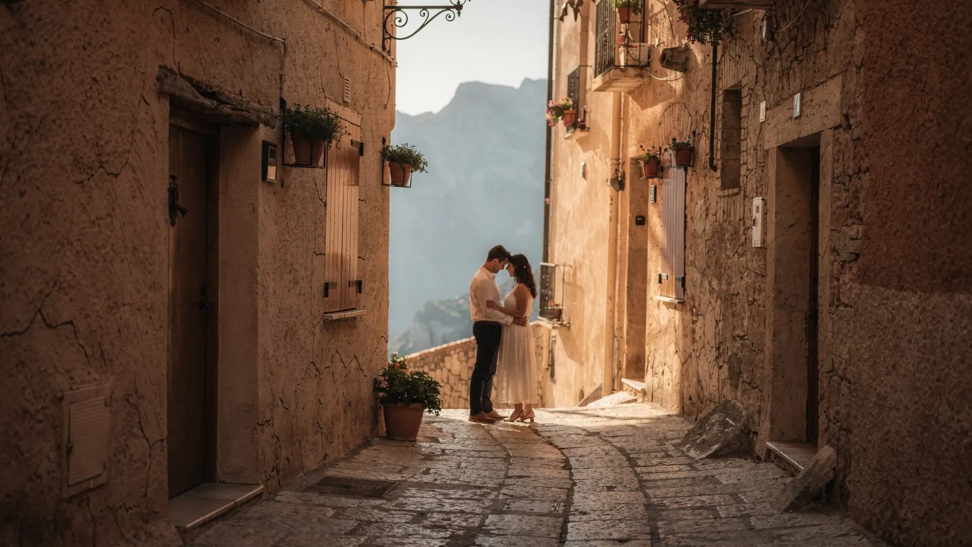 A narrow stone street in a tiny Spanish mountain village with warm textured walls, simple wooden doors, and a curve leading toward a glimpse of distant peaks. A couple stands close in golden hour light, framed naturally by the alleyway, with a quiet, intimate mood.