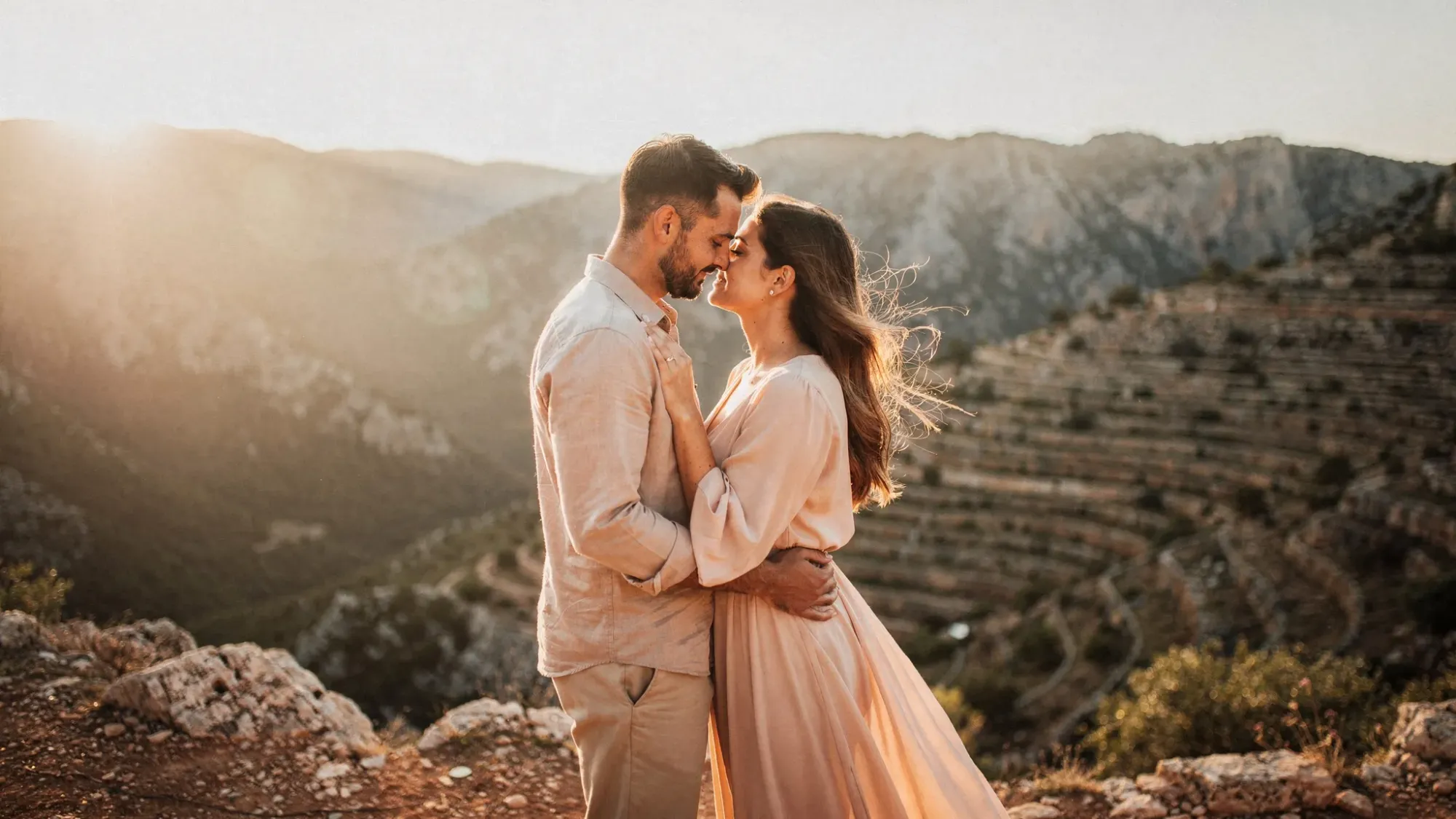 A couple standing close together on a rocky hillside near Confrides at golden hour, with soft wind moving through their clothes, Alicante mountain ridges behind them, and warm light touching the terraces below.