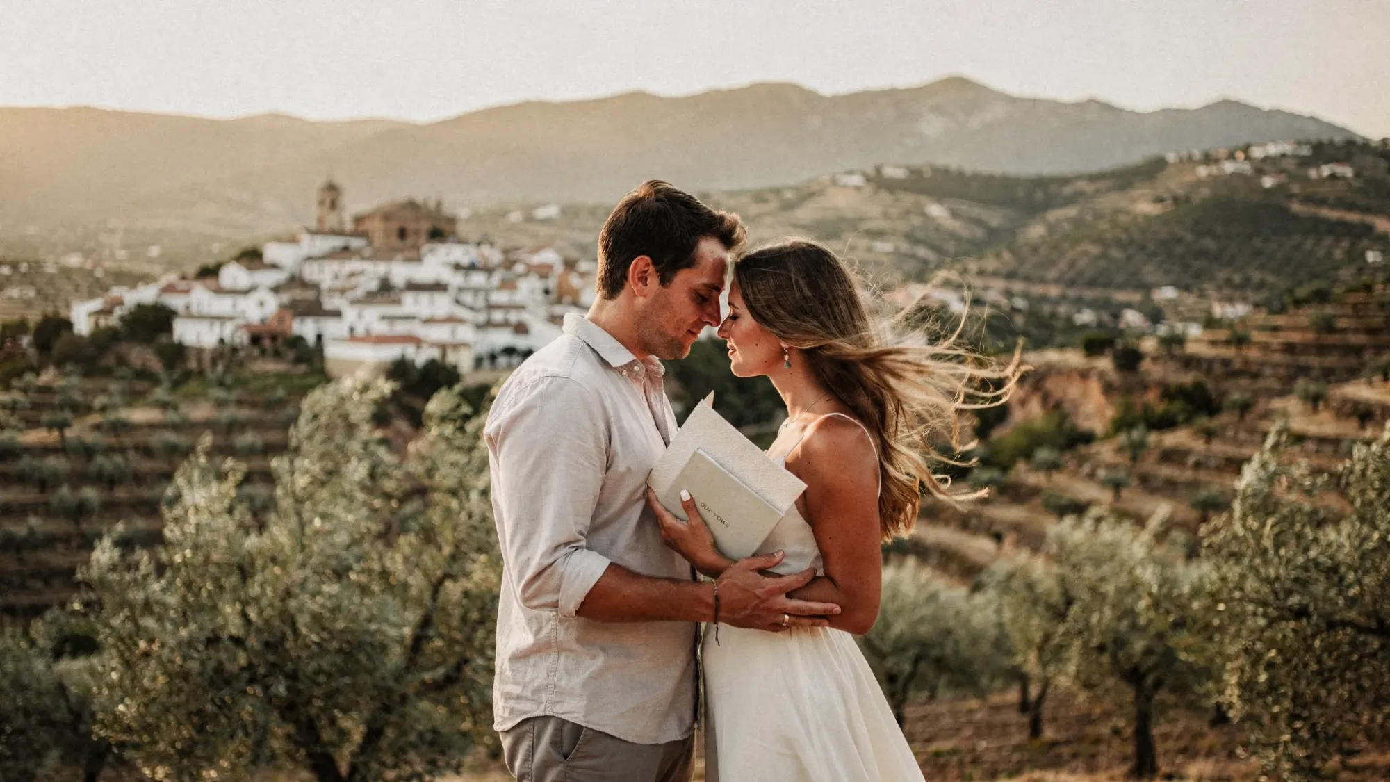 A couple stands together on a quiet hillside near an inland Spanish village, with olive trees below, soft golden light on the mountains, and no guests or decorations except simple vow books.