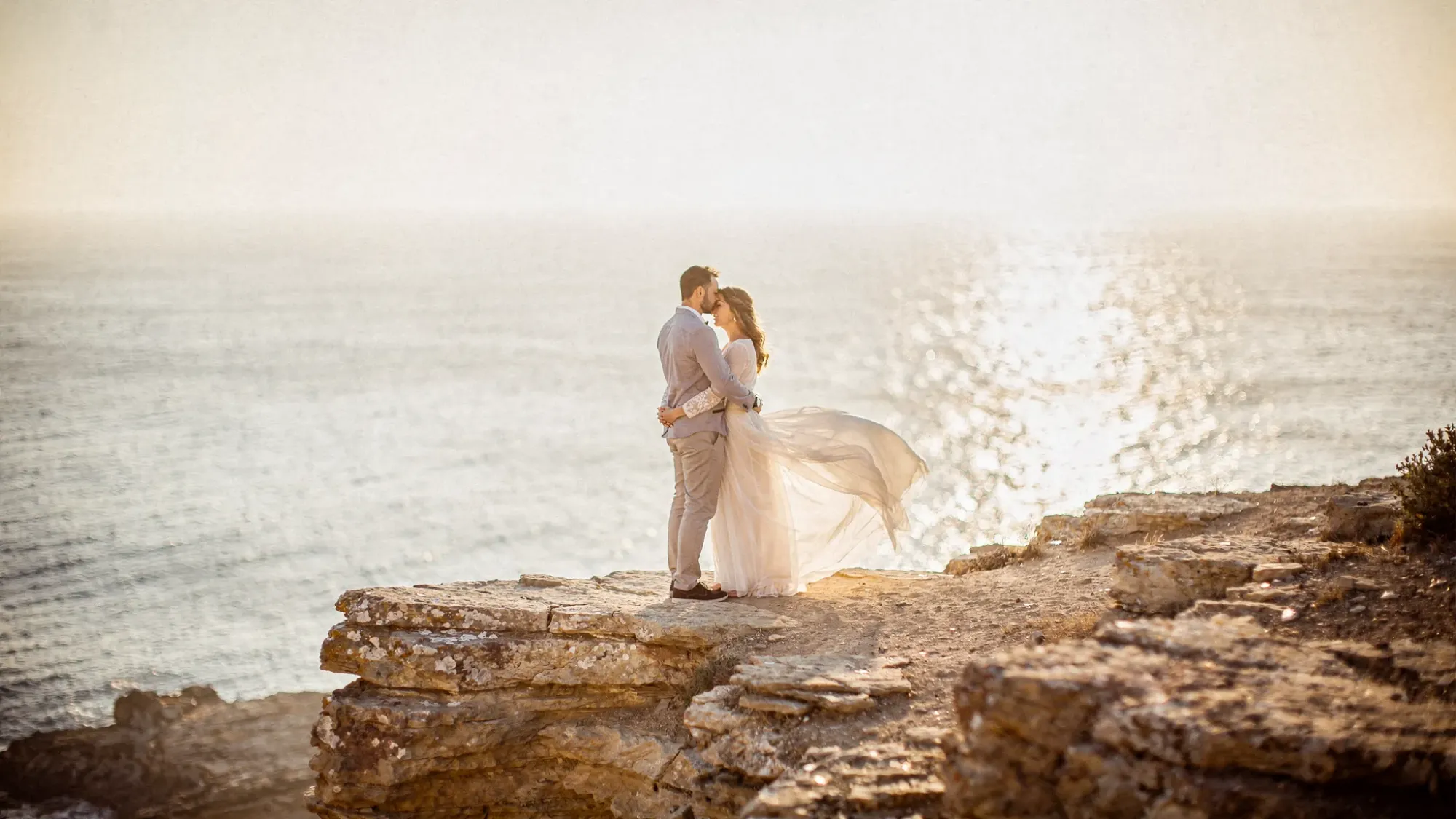 A cliffside Mediterranean elopement at golden hour, with warm light hitting textured rock, wind moving through the couple’s clothing, and the sea far below, creating a quiet cinematic mood.