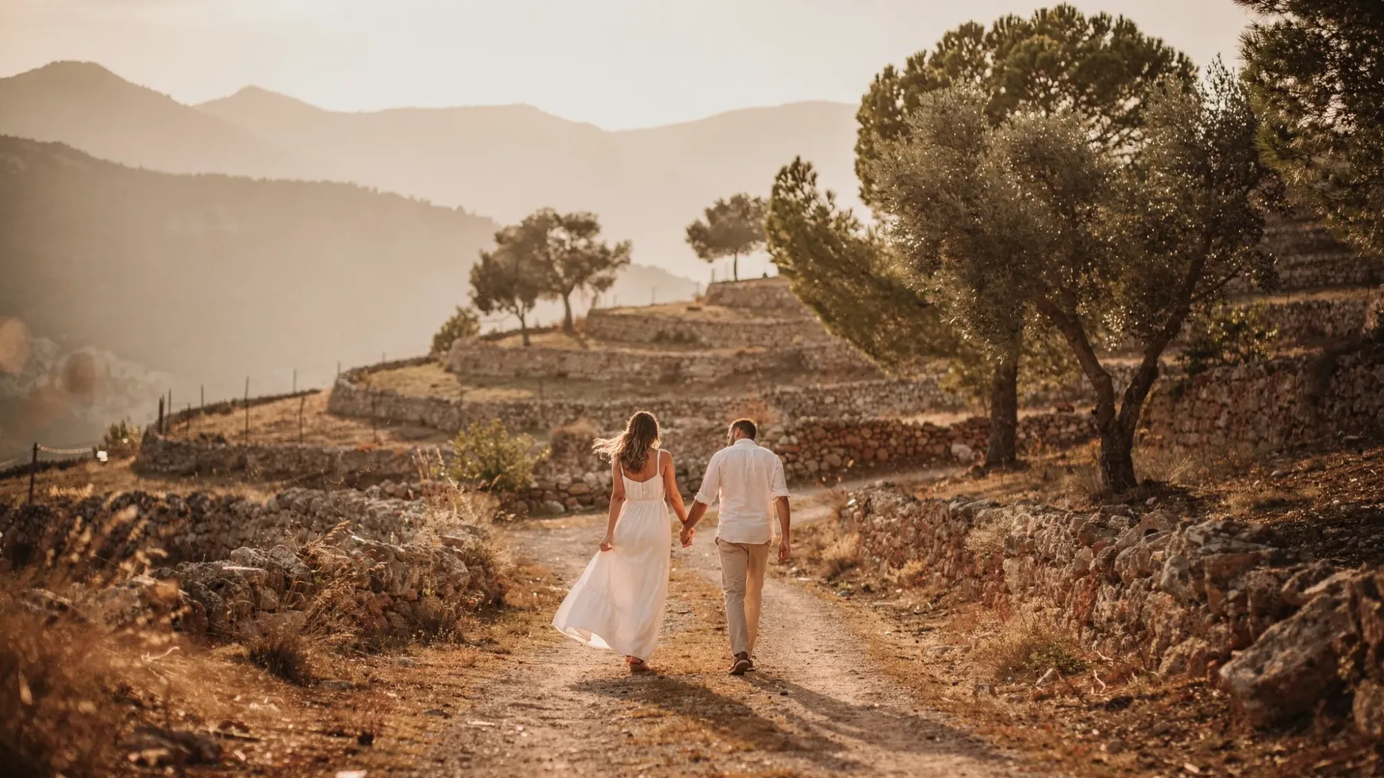 An intimate elopement scene in the mountains near Alcoleja at golden hour, with olive terraces, pine shadows, soft amber light, and a couple walking hand in hand along a quiet path.