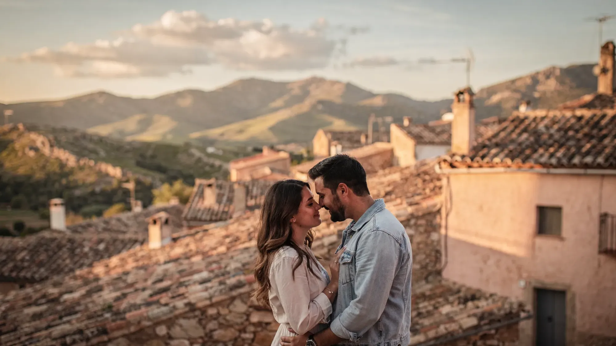A couple standing close together at the edge of a small inland Alicante village, with Penàguila stone rooftops behind them, mountain hills in the distance, and warm golden hour light on the landscape.