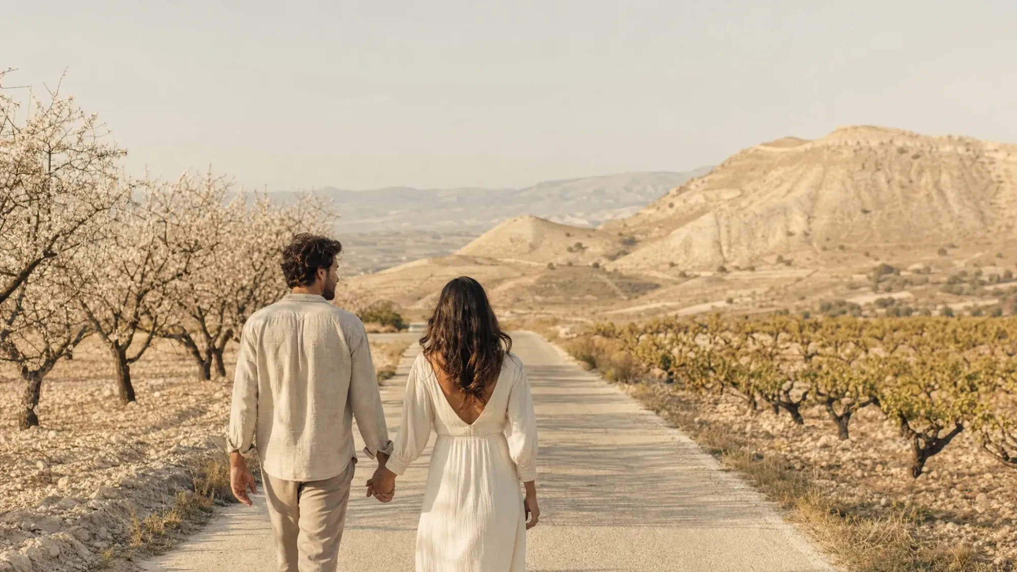 A couple walking hand in hand along a quiet country road near Algueña, with almond trees and low vineyards beside them and pale dry hills opening ahead in soft morning light.