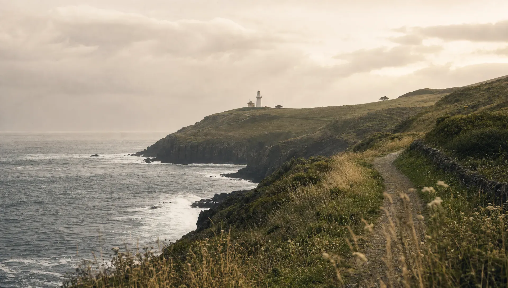 A windswept northern Spanish coastline with dark cliffs, green hills, white waves, and a small lighthouse in the distance, seen from a low coastal path under a soft grey sky.